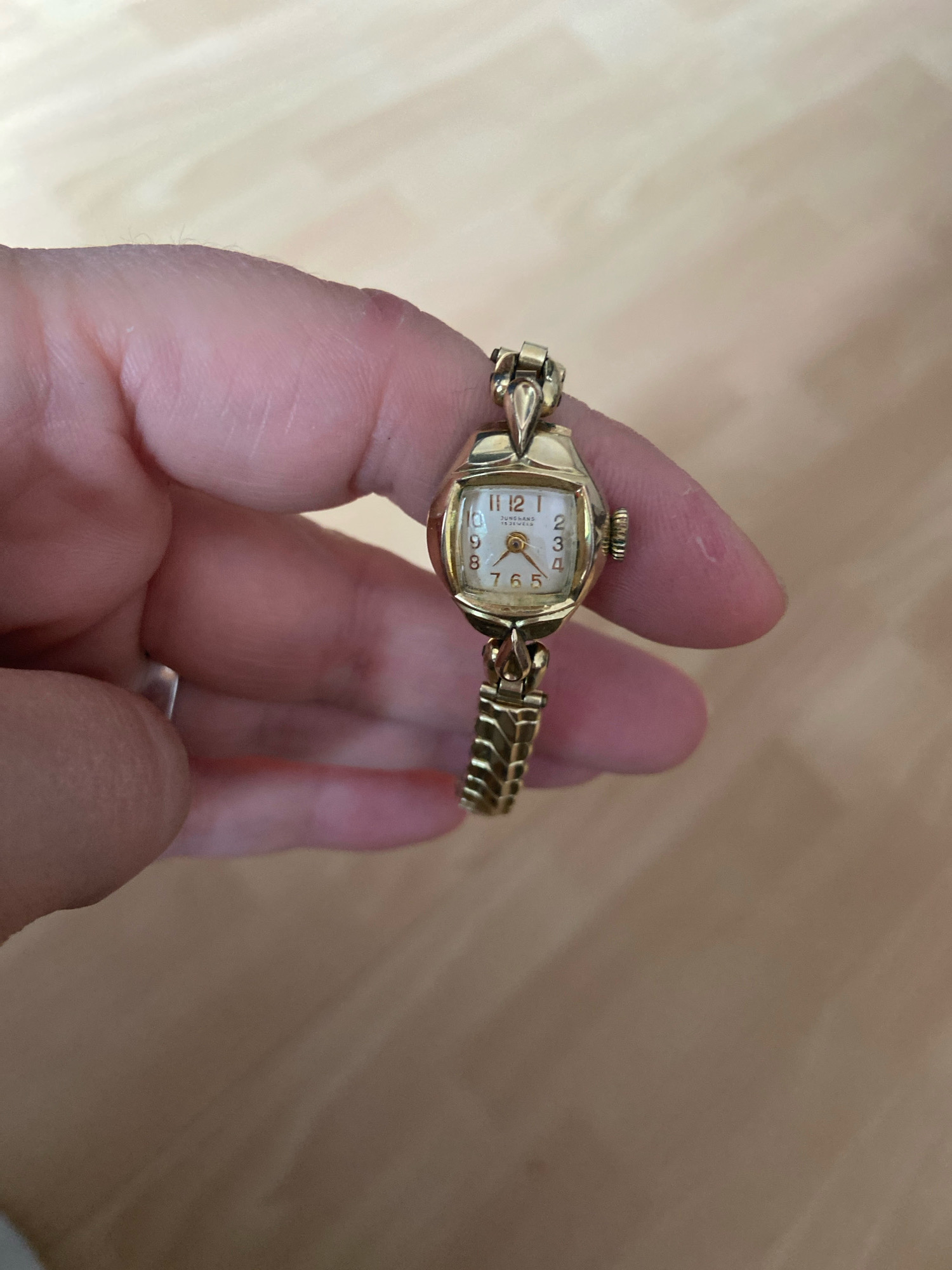 A person's hand, with visible skin texture and nails, is holding a vintage gold-colored wristwatch. The watch has a rectangular face with a white dial, black Arabic numerals, and a gold-colored hour and minute hand. The brand name "Junghans" and "17 JEWELS" are visible on the dial. The watch band is also gold-colored and appears to be made of interlocking links. The background is a light-colored wooden surface, likely a table or floor. There is no indication of the city of Kassel, Germany, in this image.