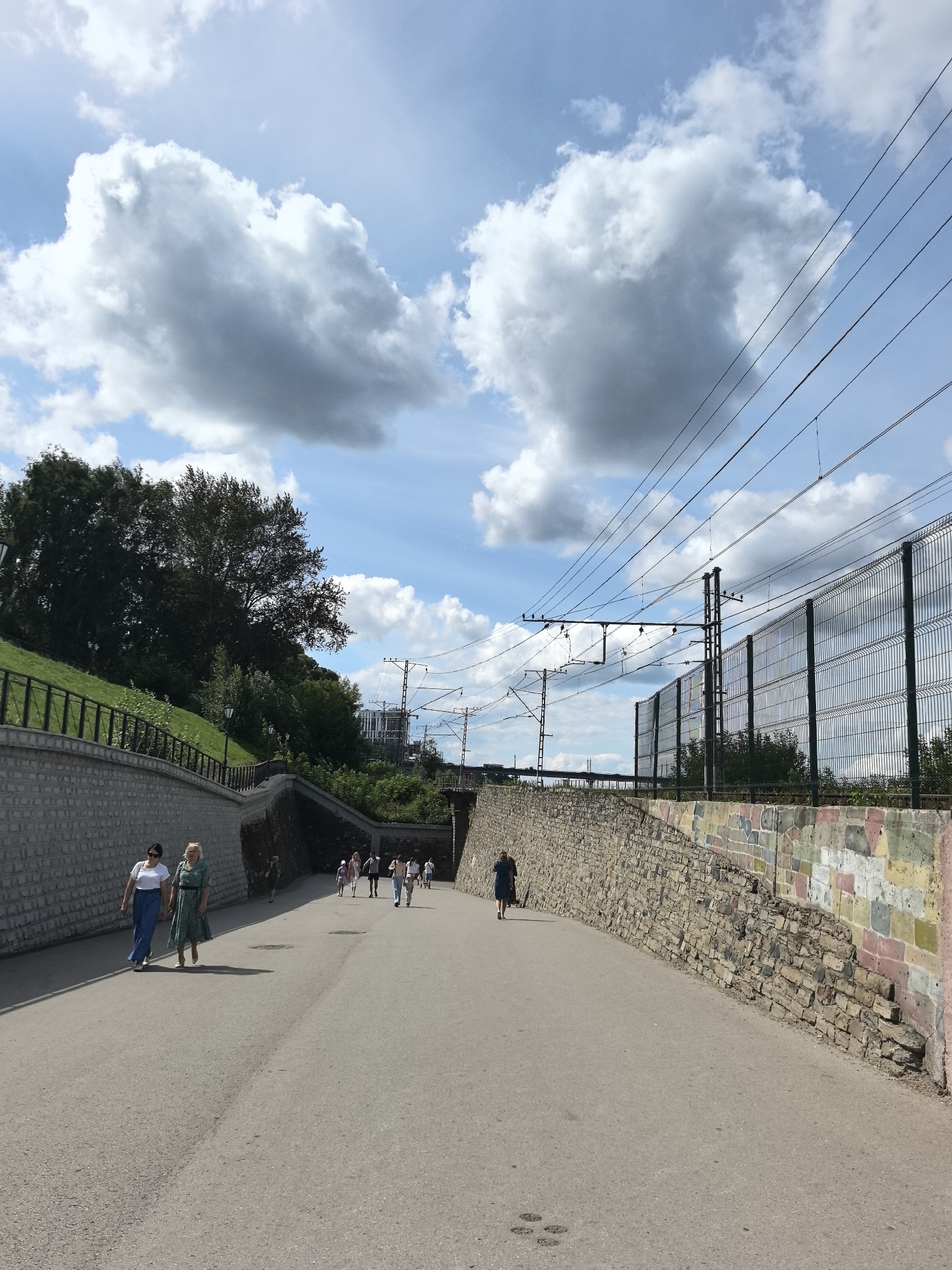 A paved pathway curves towards a tunnel entrance under a bright blue sky with large, fluffy white clouds. On the left, a stone wall with a wrought-iron fence borders a grassy slope dotted with trees. On the right, a similar stone wall, embellished with colorful rectangular blocks, runs alongside the path, eventually giving way to a wire mesh fence. Overhead, multiple sets of electrical wires for a train are strung between poles. Several people are walking in both directions on the pathway. Two women in casual attire are walking away from the camera on the left side of the path, while a group of people walks towards the tunnel entrance on the right. The scene appears to be an outdoor pedestrian area in the city of Perm, Russia, likely near a railway line.