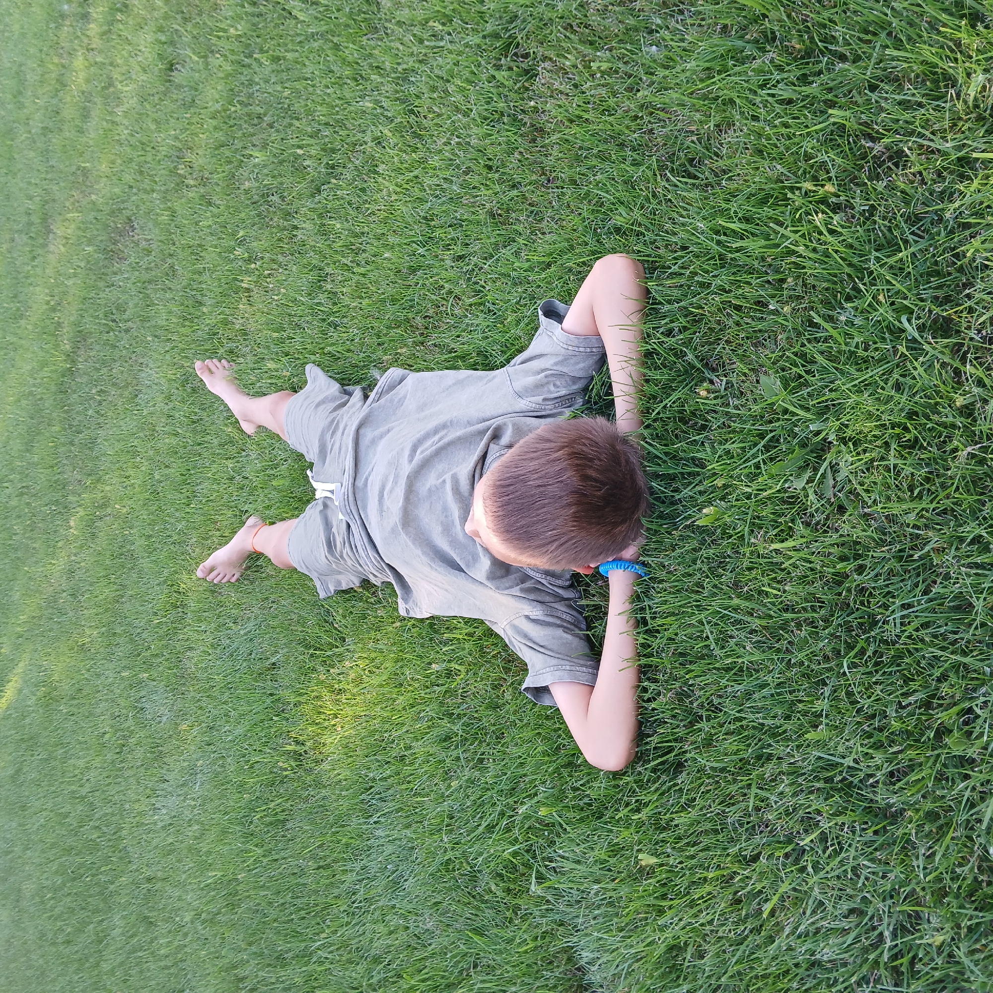 A young boy is lying on his back in a lush green grassy area. He is wearing a grey t-shirt and grey shorts. His arms are resting behind his head, and his legs are splayed outwards. He has a blue bracelet on his left wrist and an orange string on his right ankle. The grass is neatly trimmed, and there are some patches of sunlight filtering through. The scene appears to be outdoors in a park or backyard. There is no indication of the specific location in the city of Samarsky, Russia.