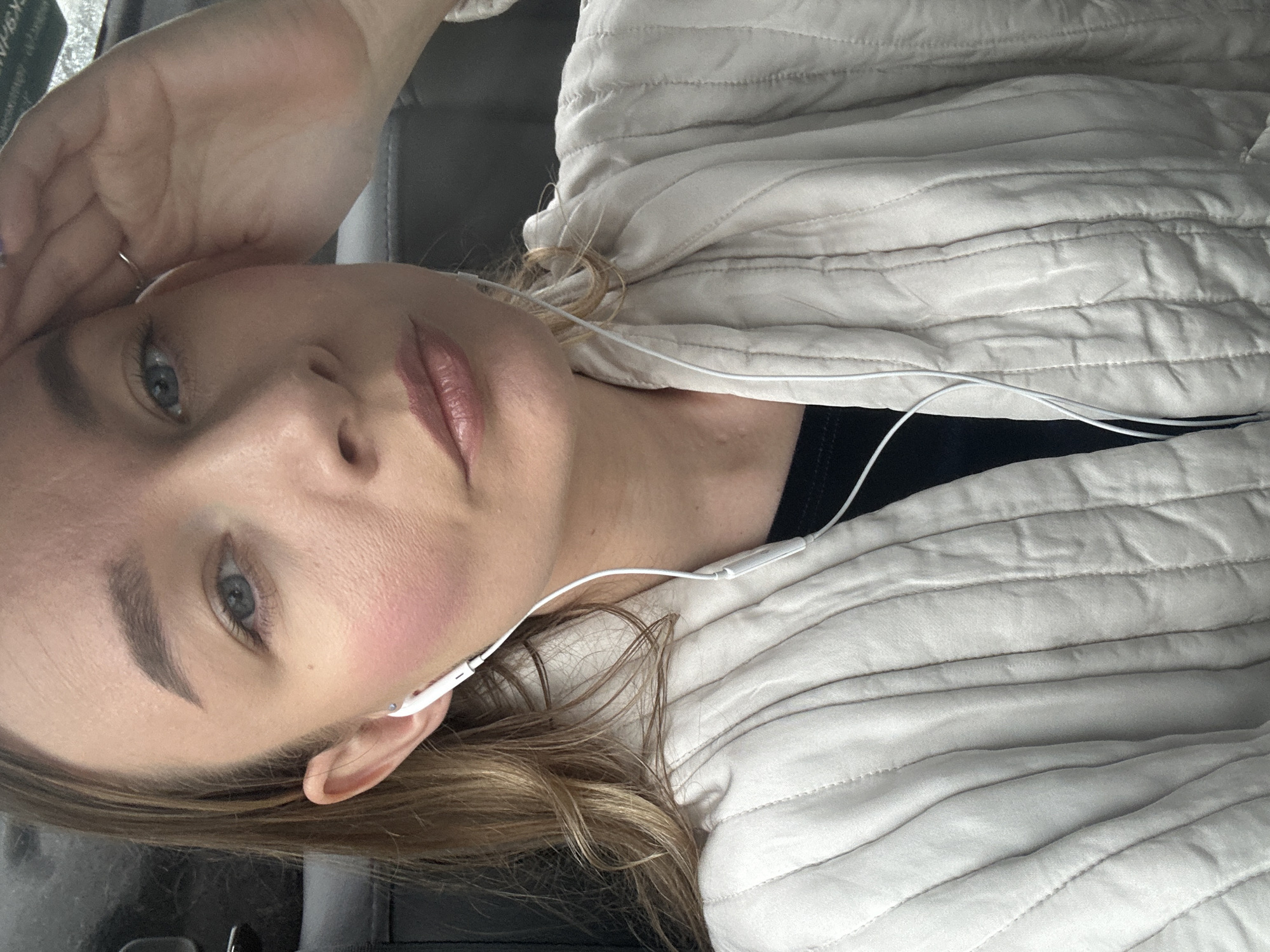 A medium shot captures a woman from the chest up, in a close-up, as she leans her head back and rests her right hand on her forehead. She is wearing a light-colored quilted jacket over a dark t-shirt and white earbuds. Her blonde hair is pulled back, with strands falling around her face and shoulders. Her skin is smooth, with a subtle pink blush on her cheeks. Her lips are painted with a glossy pink lipstick. The background is dark and out of focus, suggesting an indoor or car setting.