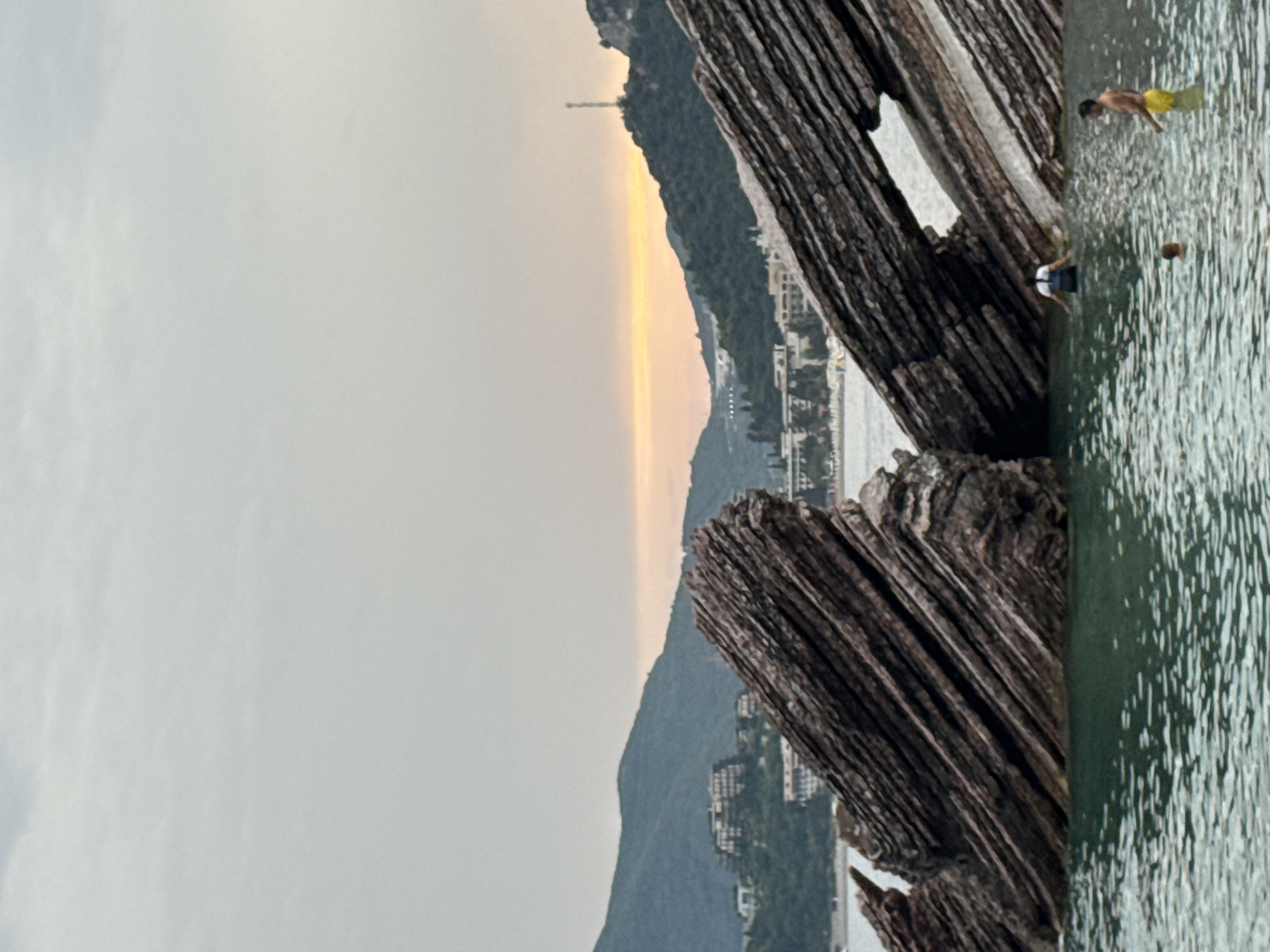 The image depicts a scenic view of the sea and coastal landscape in Rafailovići, Montenegro. In the foreground, two large, stratified rock formations jut out from the water, creating a natural arch. The water is a clear, turquoise hue, with small ripples visible on the surface.
Several individuals are present in the water. One person, wearing a white shirt and dark shorts, stands facing away from the camera. To the right, a man in yellow swim trunks stands in the water, looking towards the left. Another person, appearing to be a child, is partially submerged in the water closer to the viewer.
In the background, rolling green hills rise, dotted with buildings and structures that suggest a coastal town. The sky is overcast with a subtle hint of orange and yellow near the horizon, indicating either sunrise or sunset. A telecommunications tower is visible on one of the hilltops. The overall atmosphere is tranquil and serene, capturing a moment of relaxation by the sea.