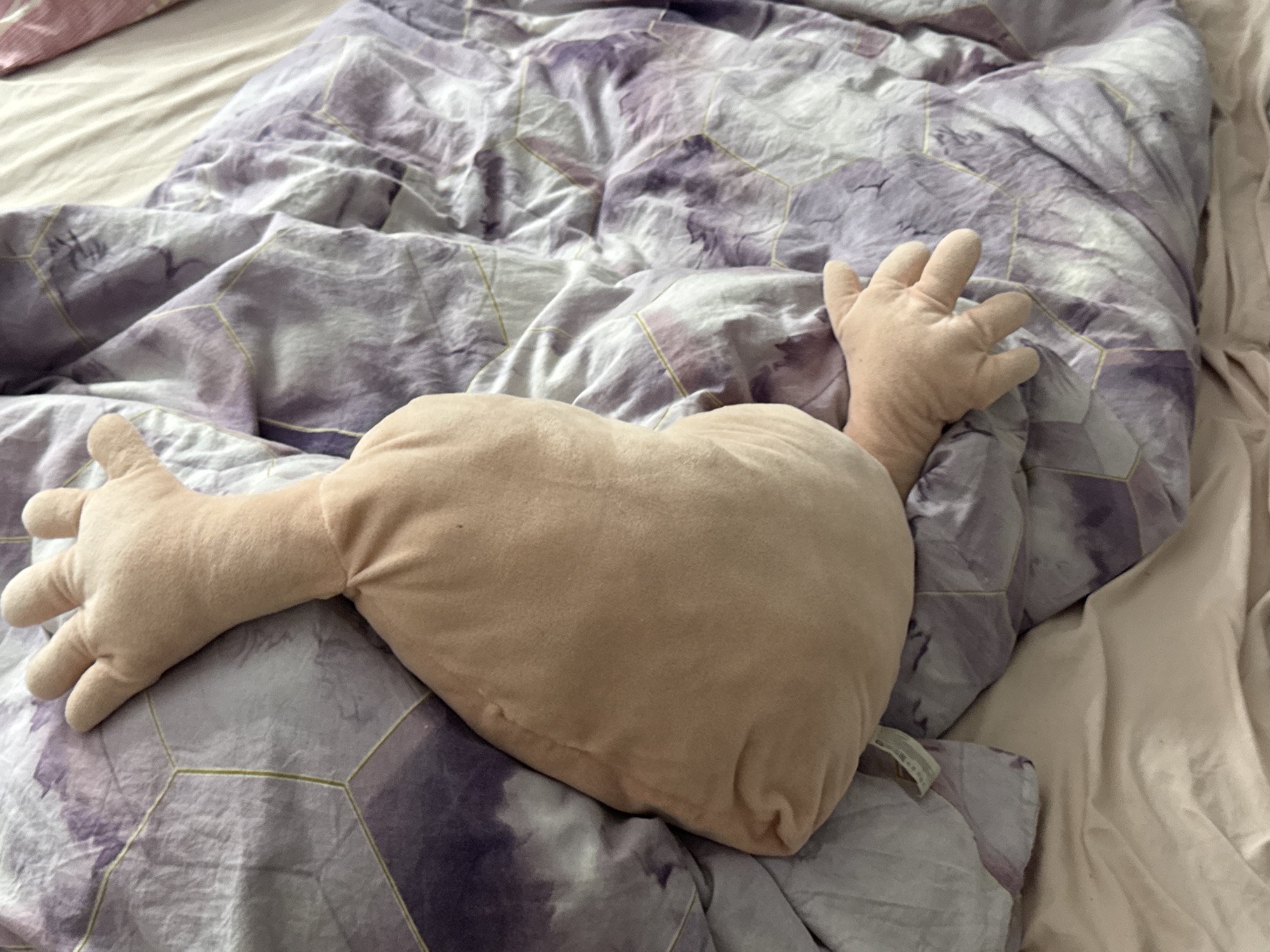 An overhead, slightly angled medium shot captures a plush pillow resembling a human-like torso with attached arms, resting on a bed with a purple and white patterned duvet. The pillow is a light beige color and is positioned centrally in the frame. Its two arms are splayed outwards. One arm is extended towards the left side of the frame, with its hand fully visible, fingers spread. The other arm is extended towards the right, also with its hand visible. The duvet features a geometric pattern with purple and white watercolor-like splotches and thin yellow lines forming hexagonal shapes. To the left and right of the duvet, portions of a plain beige sheet are visible. A hint of a pink pillow or blanket is seen in the upper left corner. The lighting suggests an indoor setting, possibly a bedroom. There are no people present in the image. The location is not specified as Saratov, Russia, as there is no visual information to support this claim.