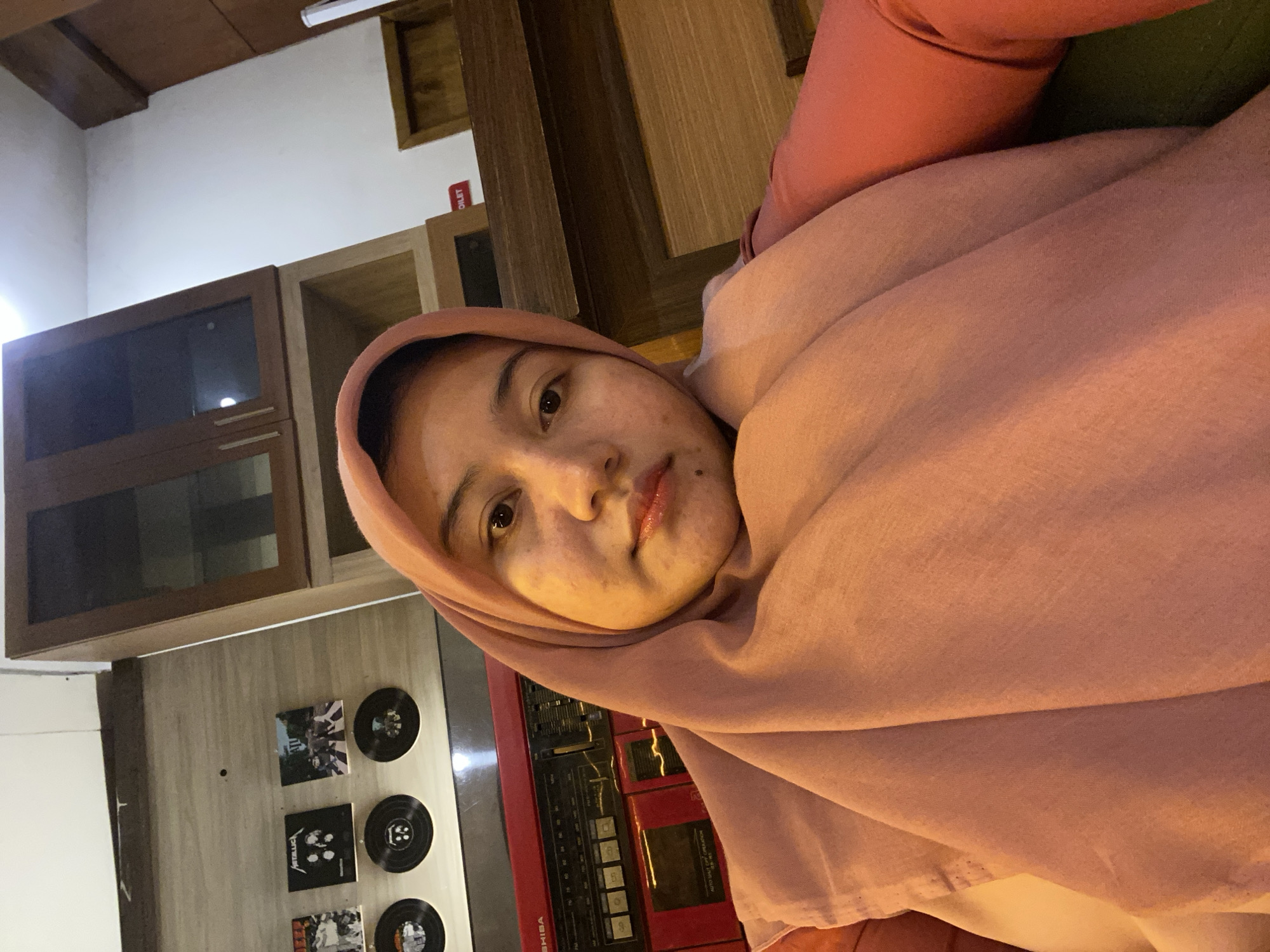 A young woman wearing a pink hijab and a pink long-sleeved shirt is taking a selfie in what appears to be a cafe or a home setting in Banjarbaru, Indonesia. The woman is in the foreground, her face is partially obscured by the hijab. Behind her, a red Toshiba boombox is visible, and above it, there are several small vinyl record covers displayed on the wall. In the background, there are wooden cabinets and a white wall. A sign indicating "TOILET" is also visible on a wooden surface. The lighting suggests an indoor environment, possibly with warm ambient light.