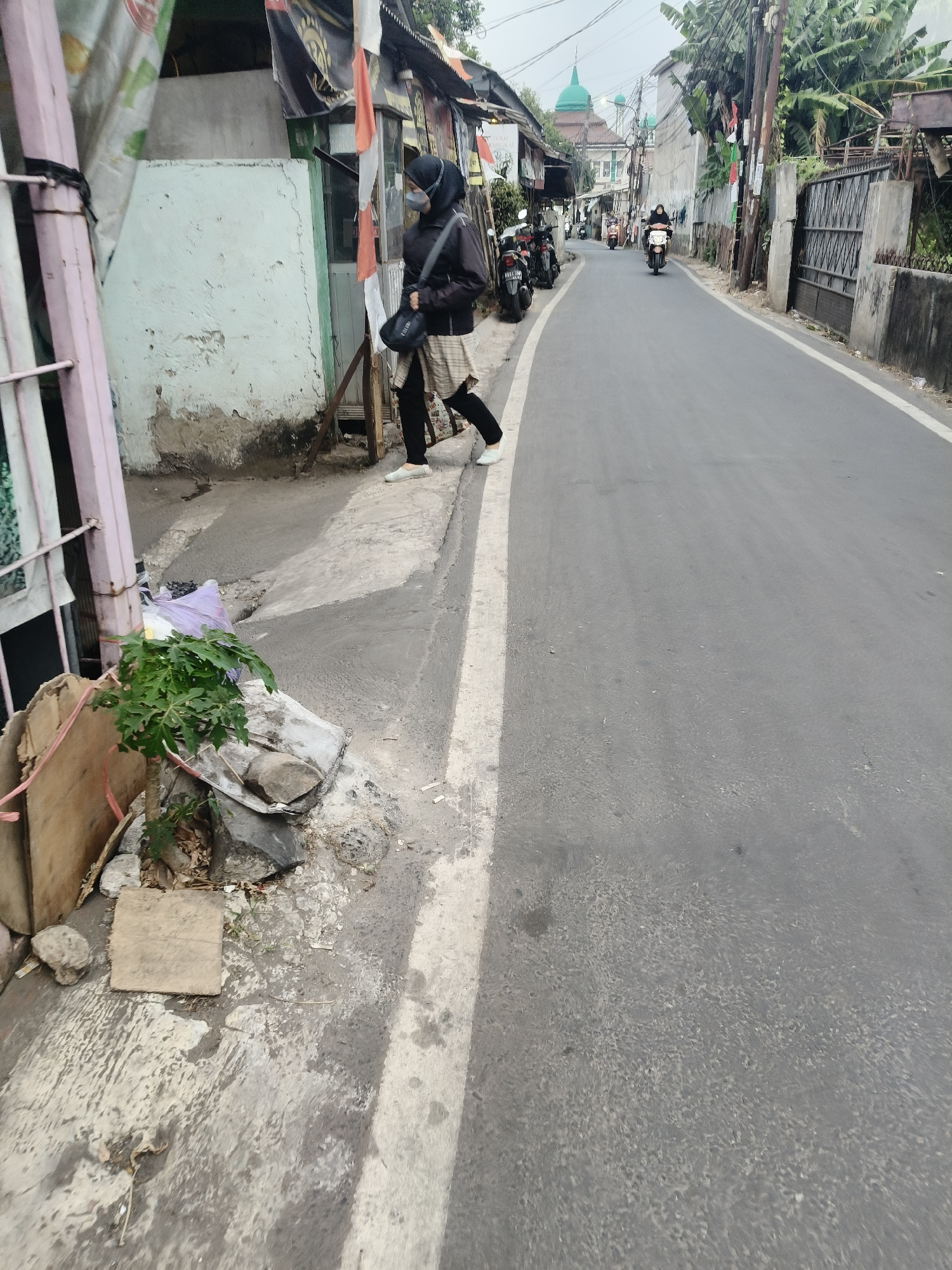 A street scene in Pamulang Barat, South Tangerang, Indonesia, features a person walking on a sidewalk next to a road. The person, wearing a black jacket, headscarf, and checkered skirt over black pants, carries a black bag. They are on a sidewalk with loose debris and a small plant. The road is paved and appears to be a two-way street with several motorcycles in the distance, one actively being ridden. Buildings line the right side of the road, some appearing to be shops with awnings. A green-domed building, possibly a mosque, is visible further down the street. The environment is urban, with a mix of modest structures and greenery.
