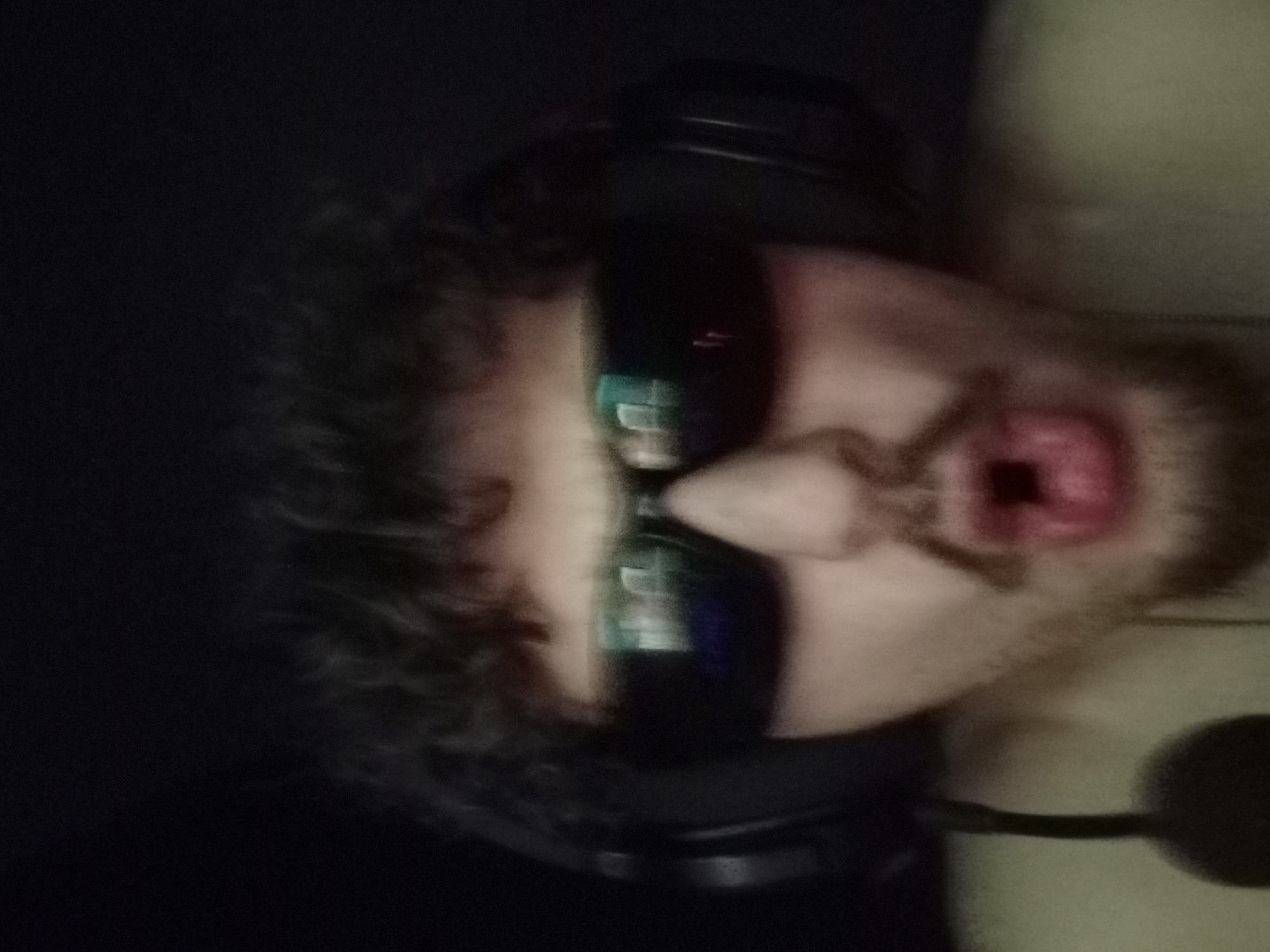 The image shows a close-up, slightly blurry, headshot of a person wearing dark sunglasses and headphones. The person has curly brown hair and is looking directly at the camera with their mouth open. The background is dark and indistinct. The subject is wearing a light-colored shirt. A microphone is visible on the left side of the frame. There is no indication of any specific location within the city of Parvomay, Bulgaria.