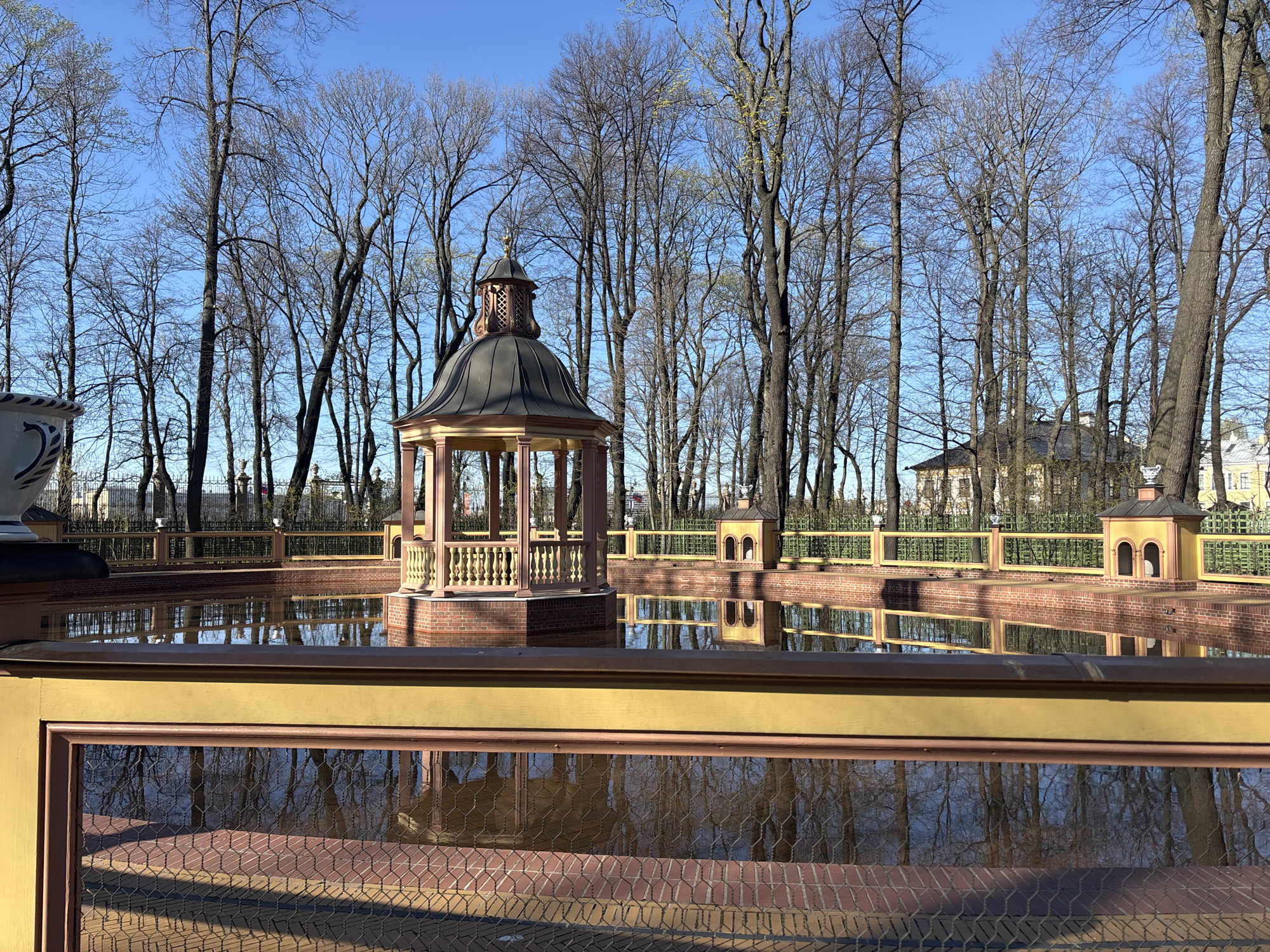 The image shows a pond in the Summer Garden in Saint Petersburg, Russia. The pond is surrounded by a low, yellow and red brick wall. In the center of the pond is a gazebo with a dark gray roof and light brown columns. Small, matching structures are located along the edge of the pond. The pond reflects the gazebo, the structures, and the surrounding trees. The trees are bare, suggesting it is early spring or late fall. In the background, there is a building with a dark roof. The sky is blue. The foreground shows a yellow, decorative barrier, possibly a fence, with red brick trim. The barrier appears to be in front of the pond.