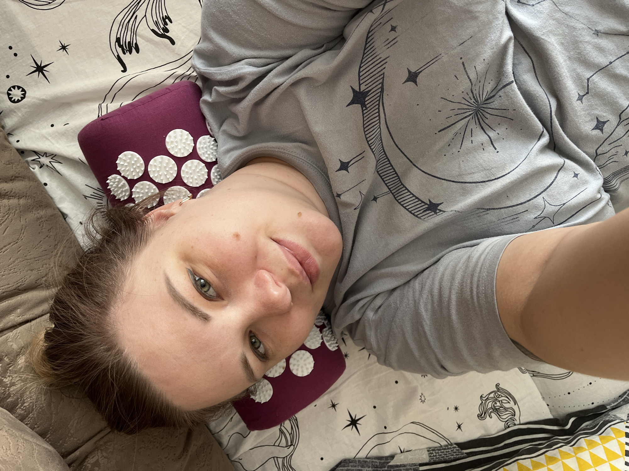 A woman with fair skin and light brown hair is lying on a bed. Her head rests on a purple pillow with white circular protrusions, and she is wearing a grey shirt with a celestial design. The bed has a patterned sheet with stars and other cosmic motifs. The location is not discernible from the image.