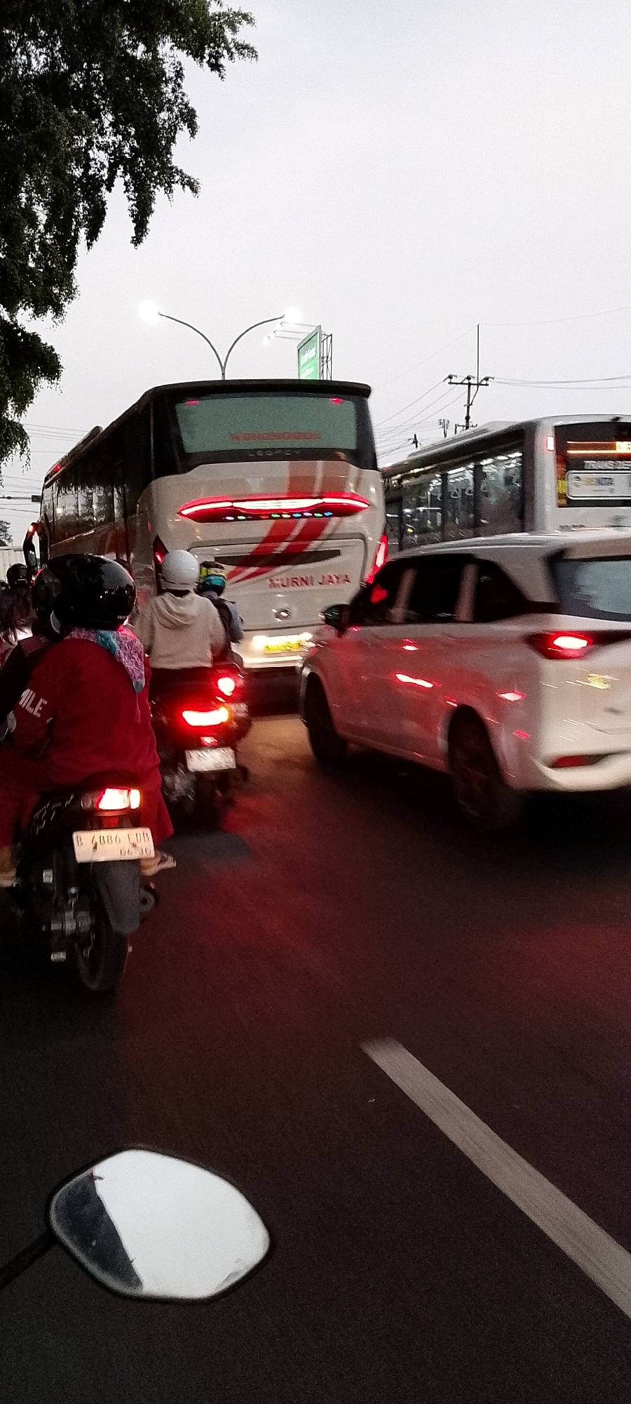 The image depicts a traffic jam on a street in Depok, Indonesia, likely on Jalan Margonda Raya, a major thoroughfare known for frequent congestion. A large white and red MURNI JAYA bus is in the center of the frame, its brake lights illuminated. To its left, a white car with its brake lights on is visible. In the foreground, a motorcycle with a rider wearing a red jacket and a black helmet is stopped. Behind this motorcycle is another motorcycle, also stopped, with a rider in a light-colored hoodie and a white helmet. A third motorcycle with a rider wearing a helmet is partially visible to the far left. The environment appears to be urban, with streetlights and power lines visible in the background. The sky is overcast. The road surface is asphalt, with a white lane marker visible in the lower right quadrant. The overall scene conveys a sense of stalled traffic and the daily commute in a busy Indonesian city.