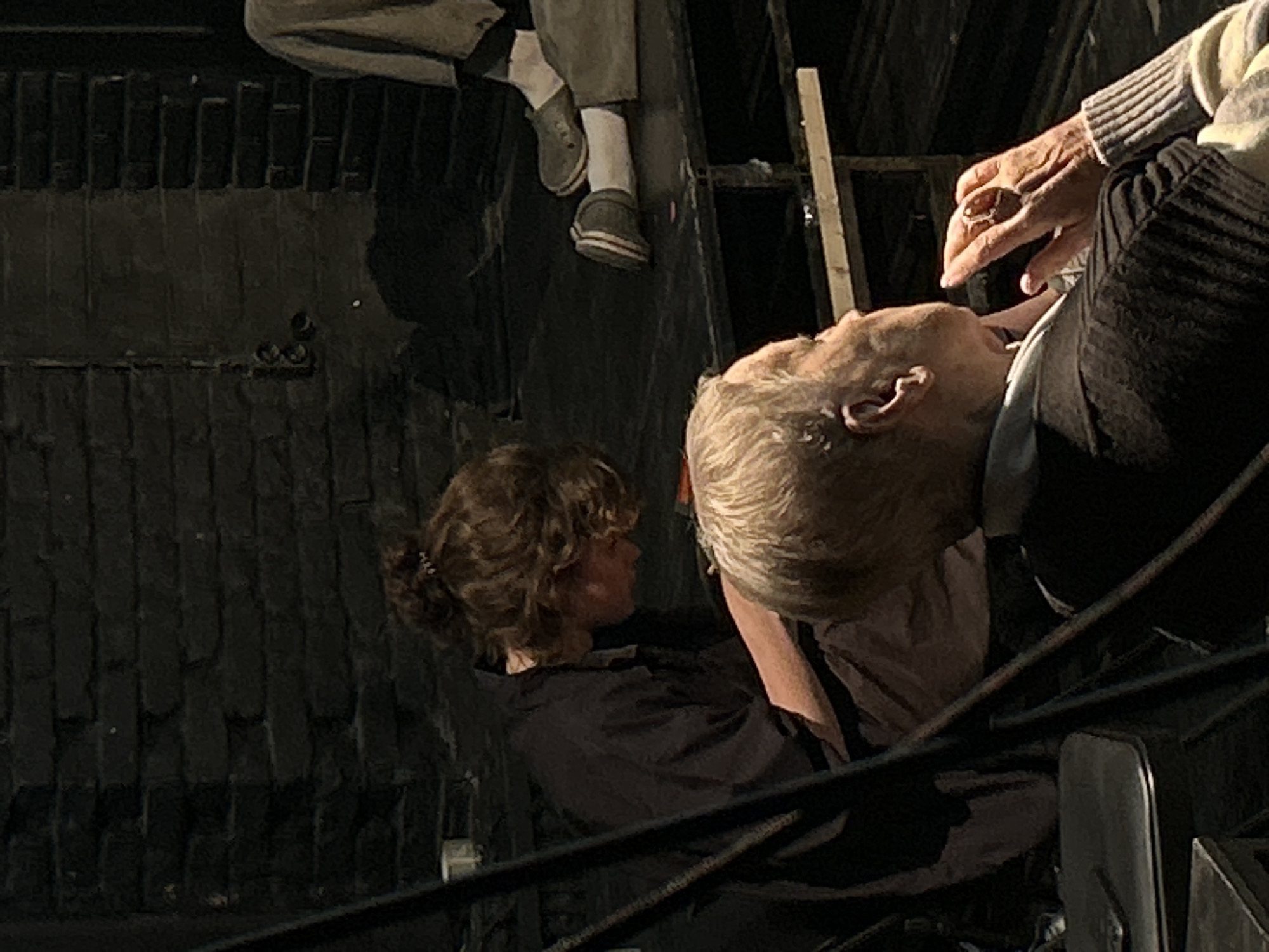 The image captures a dramatic, dimly lit scene from an elevated perspective, looking down upon several figures in an unconventional arrangement. The dominant colors are dark greys, blacks, and browns, with highlights on the subjects.
In the foreground, slightly to the right, an older person with short, light-colored hair is seen from the chest up, leaning significantly backward with their head tilted. Their right hand, appearing aged, is raised near their face, holding a small, light-colored, irregular object between their fingers. They are wearing a light-colored, possibly striped or textured sweater and dark trousers.
To the left, slightly behind and below the first person, another individual with curly brown hair tied back is visible in profile, looking towards the first person. They are wearing a dark shirt or jacket and their posture suggests they are also leaning or positioned at an unusual angle.
In the upper background, the lower legs and feet of a third figure are prominently displayed, hanging downwards. This figure wears light-colored trousers, white socks, and grey-green shoes with white soles, and appears to be a mannequin or prop due to its stiff, lifeless pose.
The background consists of dark, textured surfaces resembling a rough wall or floor with prominent vertical and horizontal grooves, possibly made of brick, stone, or patterned wood planks. Dark horizontal railings or bars are visible across the lower portion of the frame, reinforcing the impression of looking down from above. A light-colored wooden plank or beam runs vertically on the right side of the frame.
The overall scene evokes a theatrical or art installation setting, characterized by dramatic lighting and an unusual composition that creates a sense of tension or narrative. There is no visible text.