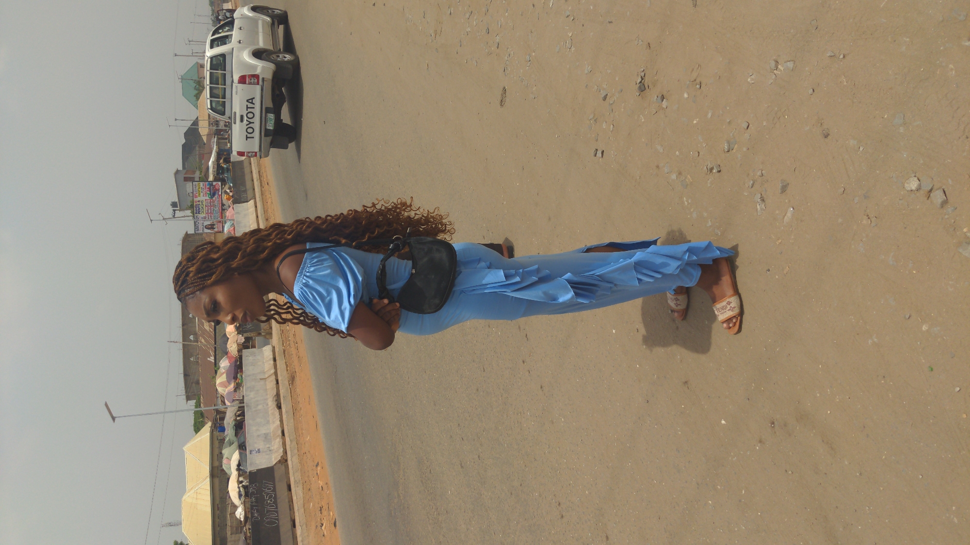 A medium shot shows a woman standing in the middle of a road in Ado, Nigeria. She has brown, braided hair and wears a light blue dress. A black purse hangs over her shoulder. The road she stands on is paved but shows signs of wear, with patches of dirt and gravel along the edges. A white Toyota pickup truck is parked partially on the shoulder of the road in the background. In the far background, there are buildings, poles, and various market stalls with umbrellas. The sky is overcast.