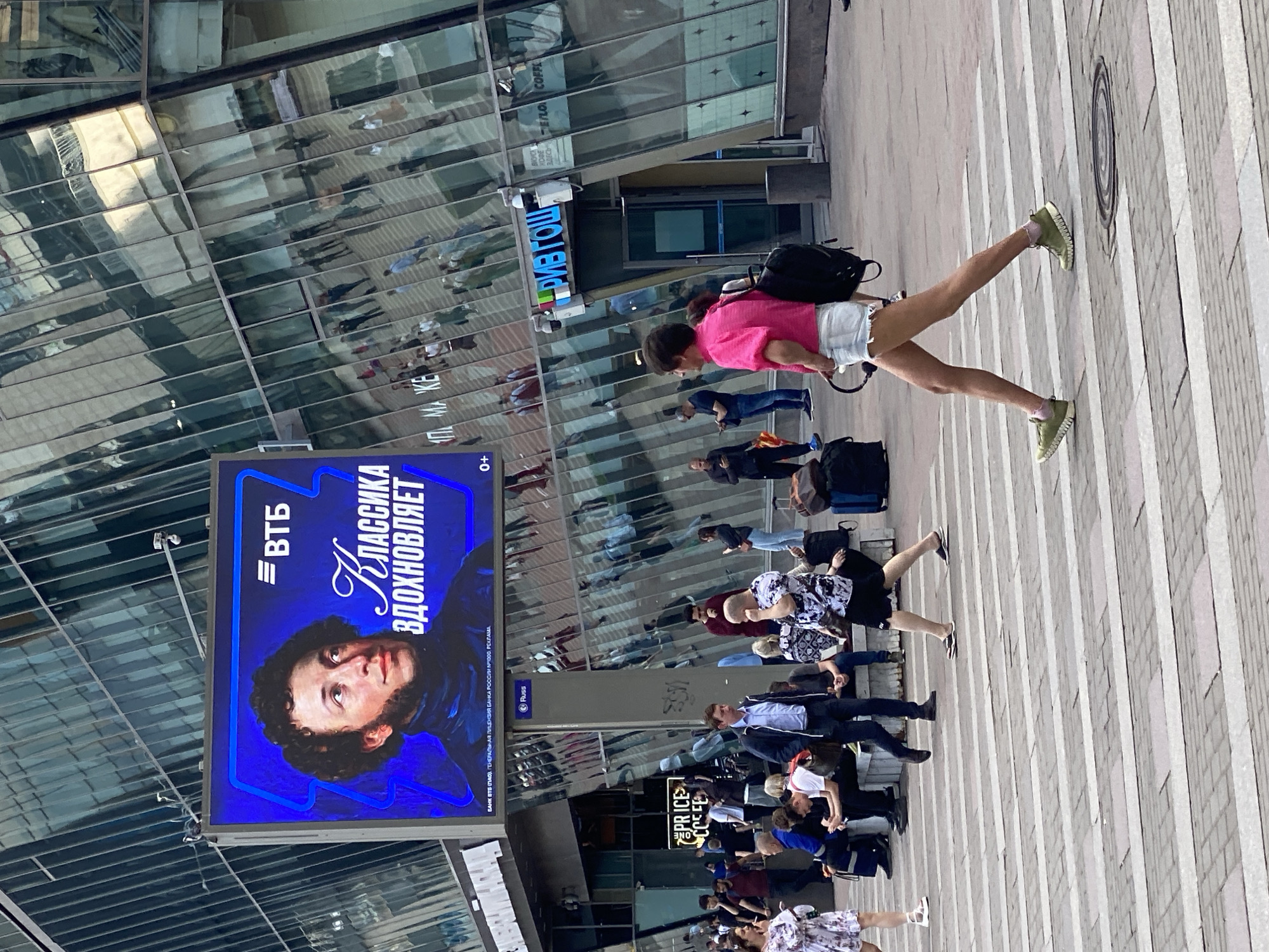 The scene, located in the city of Sinyavino, Russia, depicts a vibrant urban environment dominated by a modern glass-fronted building and a bustling pedestrian area on a bright day.
A large digital billboard prominently features a portrait of a man with curly dark hair and distinctive sideburns (identifiable as Alexander Pushkin) against a deep blue background. The billboard displays the VTB bank logo and the Cyrillic text "Классика вдохновляет" ("Classics inspire"), with smaller regulatory text below identifying the bank and its license.
In the foreground, a person with short dark hair, wearing a bright pink short-sleeved top, light blue denim shorts, and olive-green sneakers with pink ankle socks, walks confidently from right to left across a white-striped pedestrian crossing. They hold a pair of black over-ear headphones in their left hand.
Numerous other pedestrians populate the midground and background. To the left, a woman in a light floral dress and white sandals walks. Central to the midground, several individuals are seated on light-colored benches, some appearing to use mobile phones, while others stand or walk. A man in a blue button-down shirt and dark trousers, carrying a brown shoulder bag, walks towards the left. Another man in a black and white patterned shirt and dark shorts, wearing flip-flops, crosses in the opposite direction. Reflections of pedestrians and the street scene are visible across the expansive, multi-faceted glass facade of the adjacent building, creating a sense of depth and activity. Other visible figures include a man in a dark suit jacket and a man in a maroon top.
The ground is paved with rectangular light-grey pavers forming a broad pedestrian area. Street furniture includes a dark pole with a "Russ" sign, a trash receptacle, and a circular metal manhole cover embedded in the crossing. Commercial signage for "PRICE COFFEE" and "РИВ ГОШ" (RIVE GAUCHE, a cosmetics retailer) is visible on the lower levels of the buildings, along with several CCTV cameras mounted on poles and building exteriors.