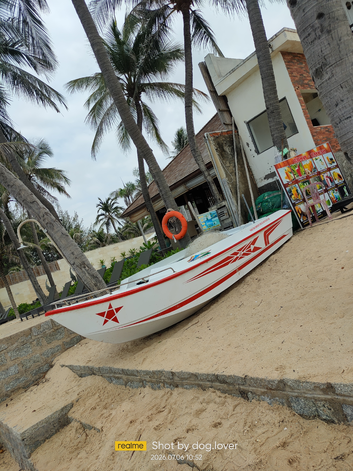 The image depicts a white speed boat with red stripe accents and a red star symbol, resting on a sandy beach. The boat is positioned on an incline leading up from the water. In the background, several palm trees tower over a low-rise building with a brick and white facade. A sign advertising juices and smoothies is visible next to the building. An orange life preserver hangs on a pole near the boat. The scene appears to be a resort or a tourist area in Phan Thiết, Vietnam. The sky is overcast, and the overall atmosphere suggests a cloudy day.