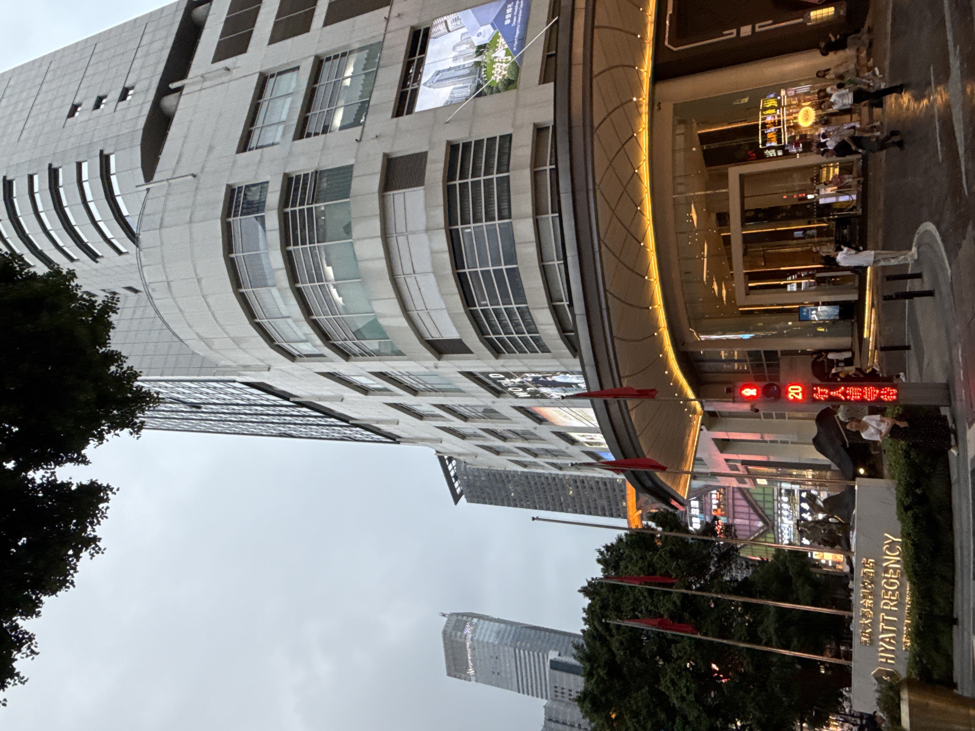 This image shows the entrance to the Hyatt Regency hotel in Yuzhong District, China, on an overcast day. The hotel is a tall, modern building with a white facade and large windows. In front of the entrance, there is a sign with "HYATT REGENCY" in gold letters. Red flags are displayed on poles to the left of the sign. The street in front of the hotel is wet, suggesting recent rain. Several people are visible on the sidewalk and crossing the street. In the background, other tall buildings can be seen, indicating an urban setting. There is also a traffic light with a red signal and the number "20" displayed on a red illuminated sign.
