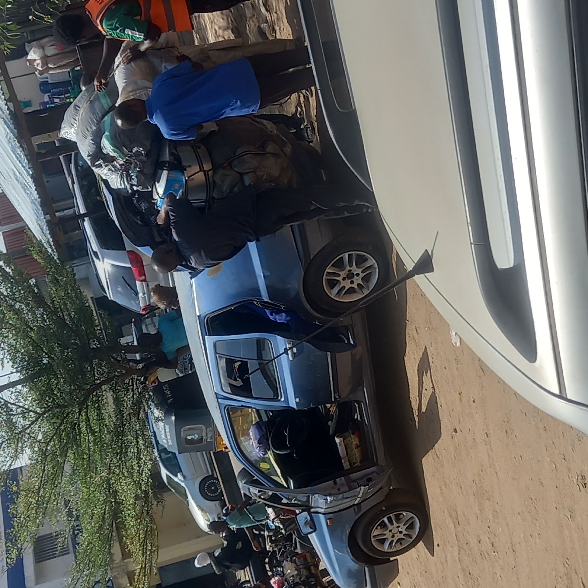 In Dadi Riba, Nigeria, a light blue Suzuki Maruti 800 is overloaded with luggage on its roof and inside the vehicle. People are loading and securing the cargo. The car's door is open. The vehicle is parked on a dirt road, among other vehicles, including a silver car and motorcycles. A building is visible in the background, as well as a tree with dense foliage. The scene suggests preparations for travel or transport of goods.