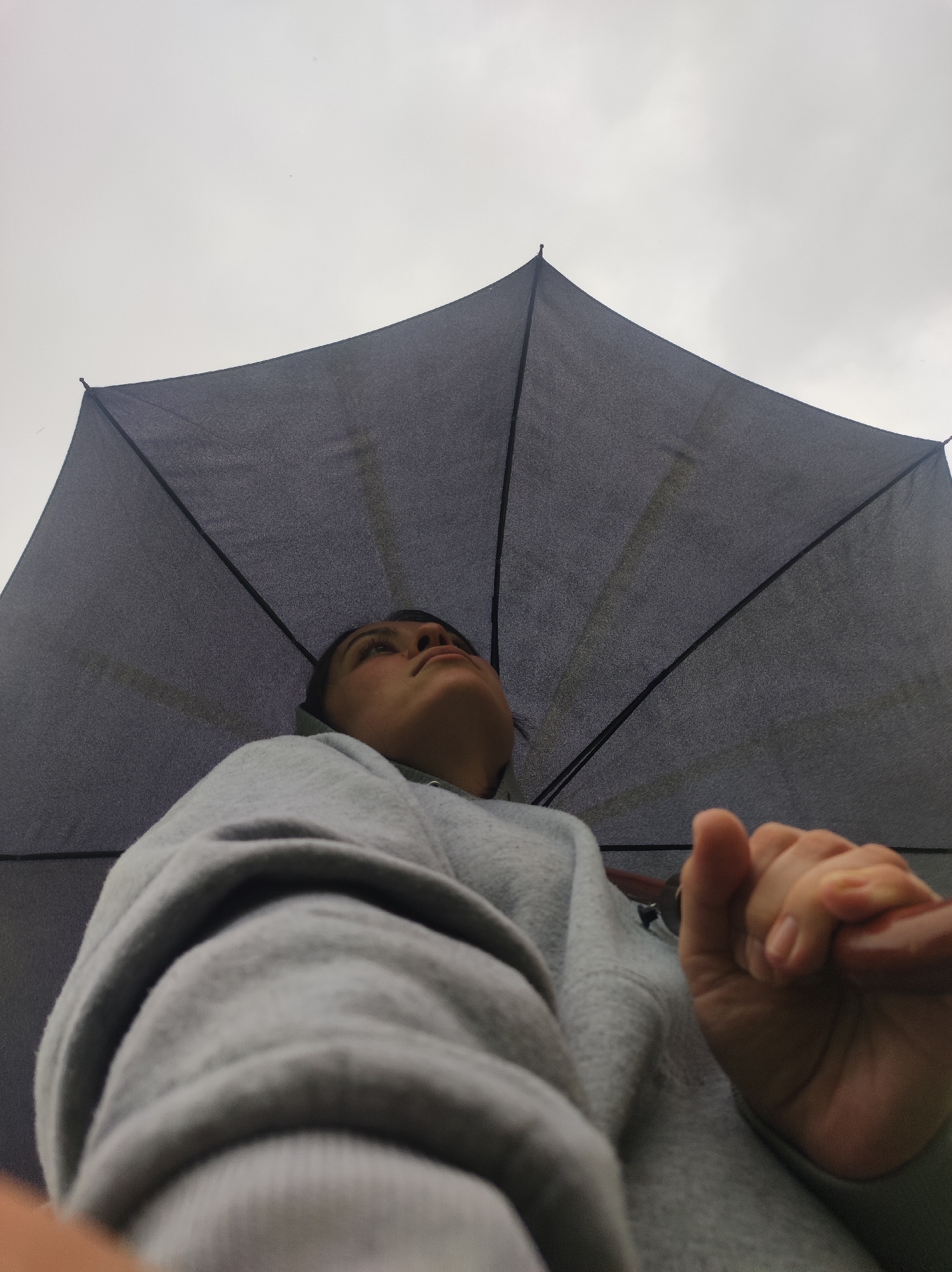 The image shows a person holding a dark grey umbrella, looking upwards towards a cloudy, overcast sky. The person is wearing a grey hooded sweatshirt. Only the person's face and hand are visible. The location is not discernible from the image.