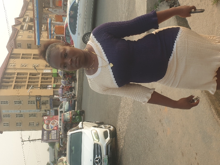 A woman stands outdoors in front of a street scene. She wears a two-toned navy and white dress and holds keys in one hand and a phone in the other. Behind her is a building with multiple windows and shops at street level, visible through traffic including cars and motorcycles. A billboard is visible, advertising a restaurant. The sky is overcast.