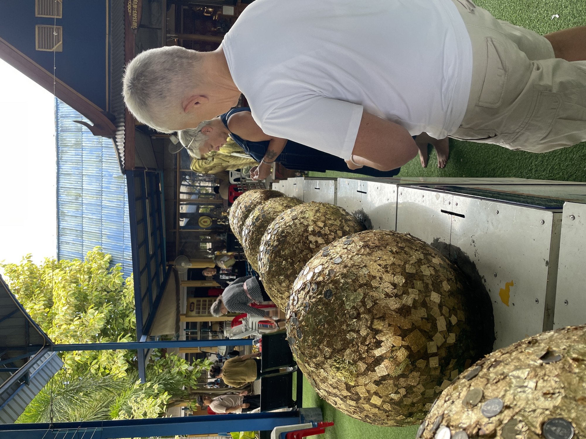 In Bang Sare, Thailand, a temple setting features a row of large, golden spheres adorned with gold leaf and coins, likely part of a Buddhist ritual. People are observing and interacting with the spheres. In the foreground, a man in a white t-shirt and beige shorts stands with his back to the camera. To his left, a woman in a dark blue jumpsuit is looking down, possibly applying gold leaf or a coin. Behind them, other individuals are visible, including a woman in a patterned shirt and a man in a light-colored t-shirt in the background. The environment includes a blue-roofed structure, lush green foliage, and artificial turf.