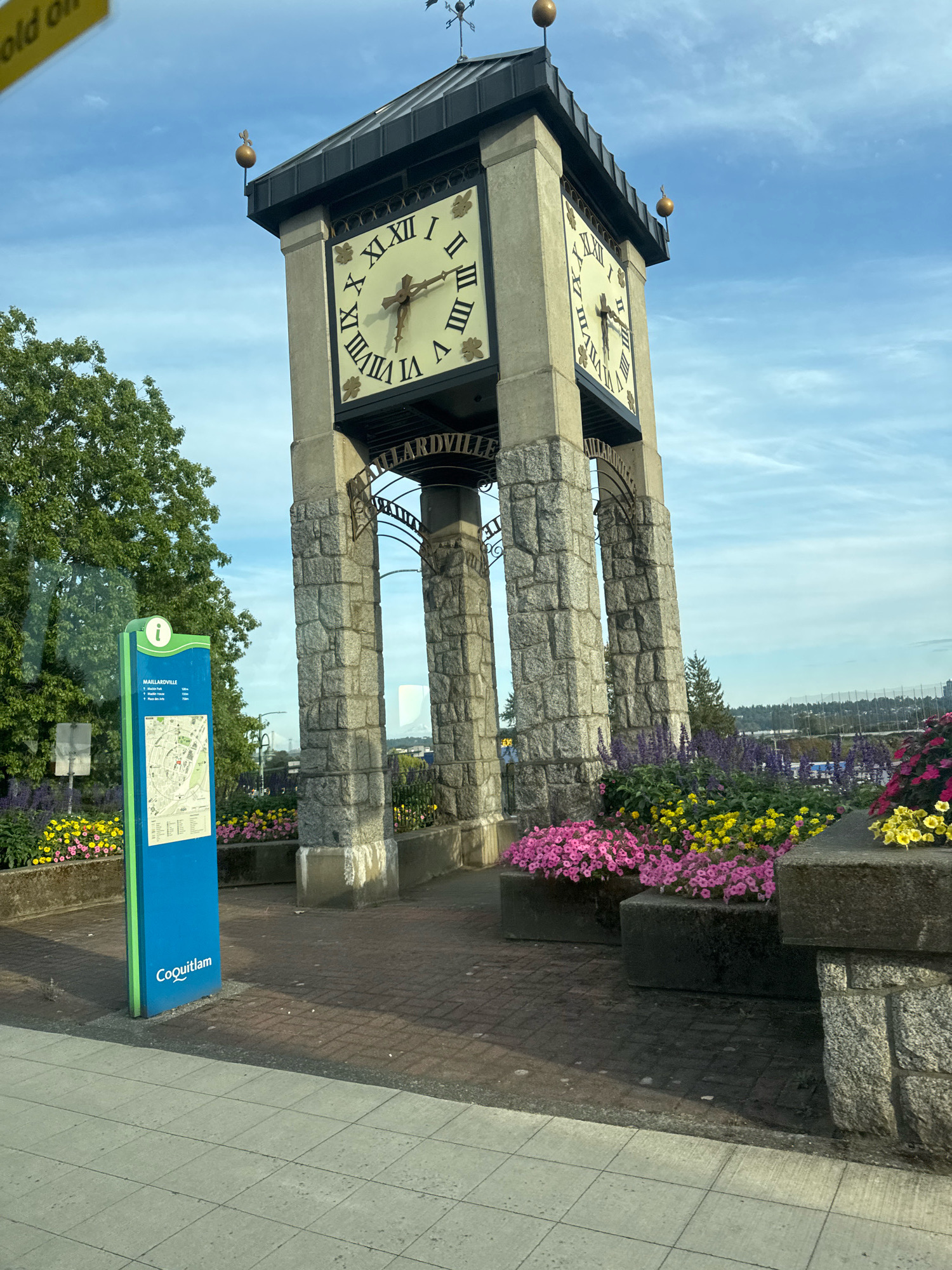 The image shows a public clock tower in Coquitlam, Canada. The clock tower is made of stone and has four clock faces with Roman numerals. There are also flowers planted around the base of the tower. A blue sign with a map of the area is visible to the left of the tower. The sky is blue with some clouds. There is no indication of any people in the image or any specific actions taking place. The environment appears to be an outdoor public space with manicured landscaping.