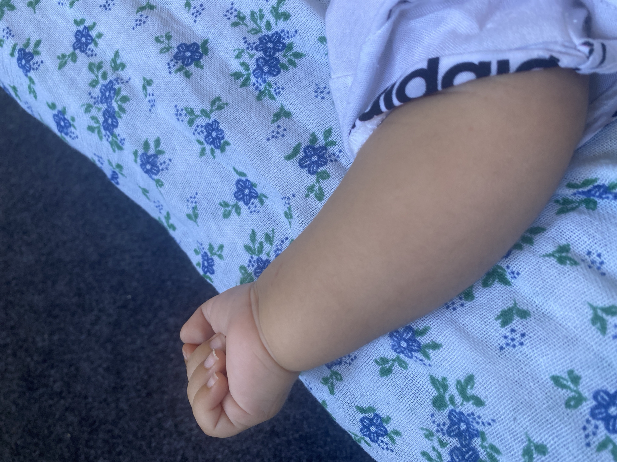 The image shows a close-up of a baby's arm resting on a light blue fabric with a floral pattern of blue flowers and green leaves. The baby's hand is visible at the top left of the frame, with fingernails clearly defined. The baby's arm is smooth and lightly tanned. A white garment with black lettering, possibly a brand logo, is visible on the right side of the arm. The background appears to be a dark, textured surface, possibly upholstery or fabric. The scene is likely indoors or in a shaded outdoor area, given the soft lighting. The image provides no information that would allow for its localization in Arbagish, Uzbekistan.