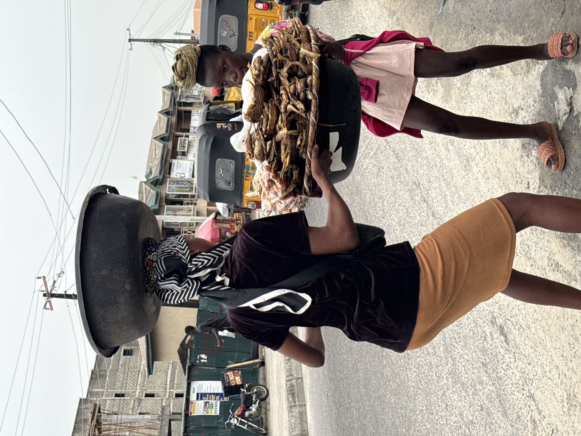 A vertical, full outdoor shot captures two African women in Ado, Nigeria, engaging in commerce on a city street. The woman on the left balances a large, dark metal basin atop her head, cushioned by a colorful patterned cloth. She wears a dark-colored top and a brown skirt, with a white-trimmed black bag slung over her shoulder. The woman on the right holds a dark container filled with dried goods, possibly fish, supported in front of her. She is dressed in a light pink dress with a red garment draped over her shoulder. Both women wear sandals and are standing on a paved road.
In the background, there is a yellow "TVS" branded vehicle and other city life elements such as buildings, utility poles, and signs. Construction or unfinished buildings are also visible. The sky appears overcast, suggesting daylight conditions.