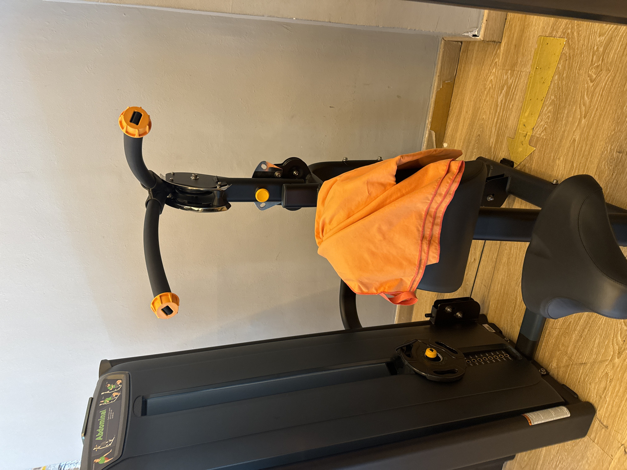 The image shows a close-up of an abdominal exercise machine in a gym. The machine is black with gray and orange accents. An orange towel is draped over the handlebars. To the right of the machine, a yellow arrow sticker points downwards on the wooden floor. The machine has a weight stack visible, and a diagram with illustrations of people performing exercises is displayed on the side. Visible text includes "Abdominal" on the diagram. The lighting suggests it is daytime, and the setting is an indoor gym. There are no people visible in the image.