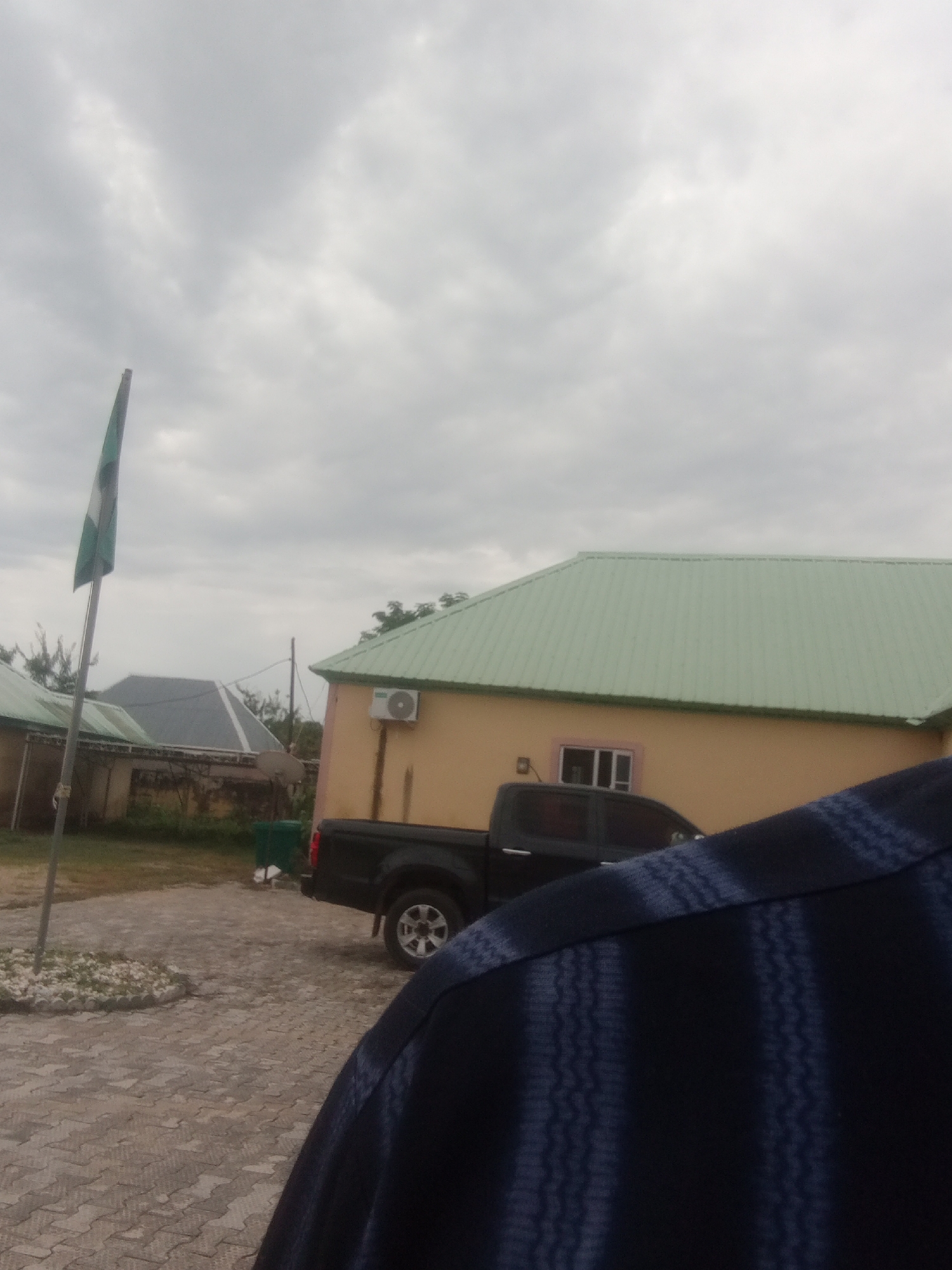 The image depicts an outdoor scene in Jalingo, Nigeria, under a cloudy sky. In the foreground, the shoulder and back of a person wearing a dark blue garment with a patterned design are visible.
The midground shows a black pickup truck parked on a paved area. Behind the truck, a light-colored building with a green roof is prominent. An air conditioning unit is mounted on the side of this building. To the left of the truck, another building with a grey, angled roof is partially visible, along with a green trash bin.
Further to the left, a flagpole stands with a Nigerian flag attached, showing green and white stripes. The flag appears to be slightly furled. The ground is covered with interlocking pavers.
The overall impression is that of an institutional or residential compound, possibly during a daytime with overcast weather conditions. There are no discernible emotions conveyed in the image, and no visible text.