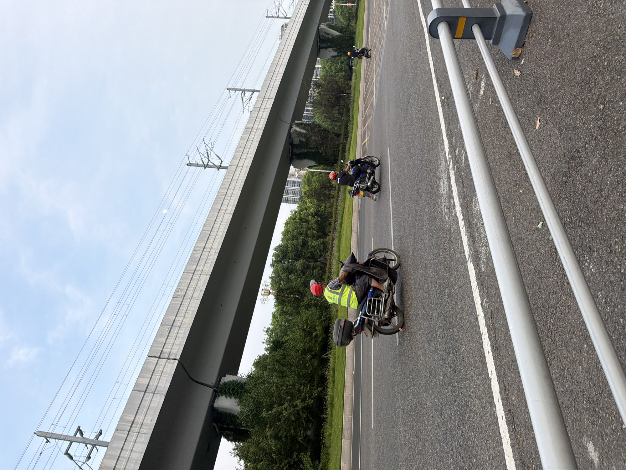 In the city of Jiangjin, China, an overhead overpass with electrical wires is visible against a hazy sky. Below the overpass, a paved road features two lanes of traffic. A man wearing a bright yellow reflective vest and a red helmet rides a blue motorcycle in the left lane. He is carrying a black storage box on the back of his motorcycle. In the right lane, another person wearing a red helmet rides a motorcycle. Further in the distance, a third motorcycle is also visible. Trees and modern buildings line the side of the road. A metal guardrail runs along the edge of the road in the foreground.