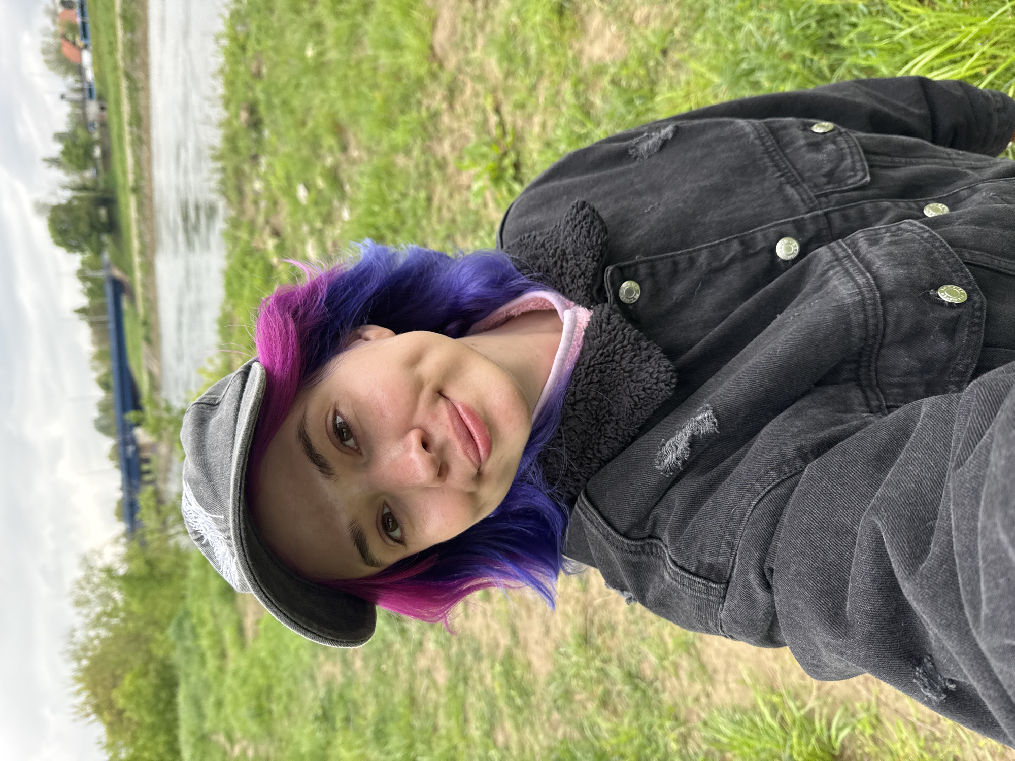The image shows a person with dyed purple and pink hair, wearing a gray cap and a dark denim jacket with a black fuzzy collar, against a background of green grass and a body of water. The person is looking at the camera with a slight smile. The location appears to be outdoors with a river or canal running in the background. It is likely in Zagreb, Croatia.