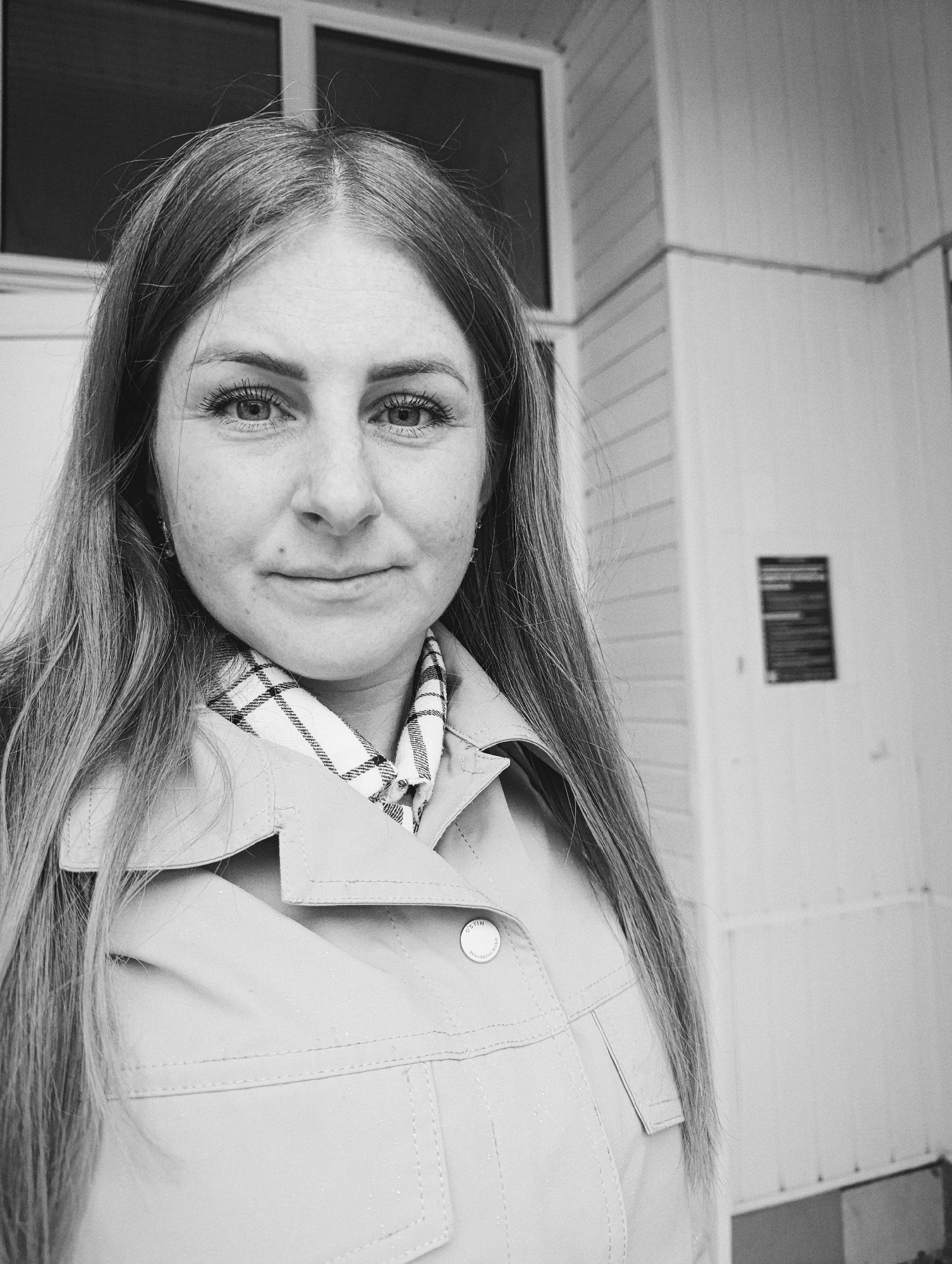 A black and white, eye-level, medium shot shows a woman looking directly at the camera with a slight smile. She has long, straight hair that is parted in the middle and falls past her shoulders. Her eyes are blue with visible eyelashes. She wears a light-colored jacket over a plaid scarf. The background is out of focus and appears to be a white building with horizontal siding and a window. There is a black sign on the wall to the right of the woman. The overall scene is clean and uncluttered.