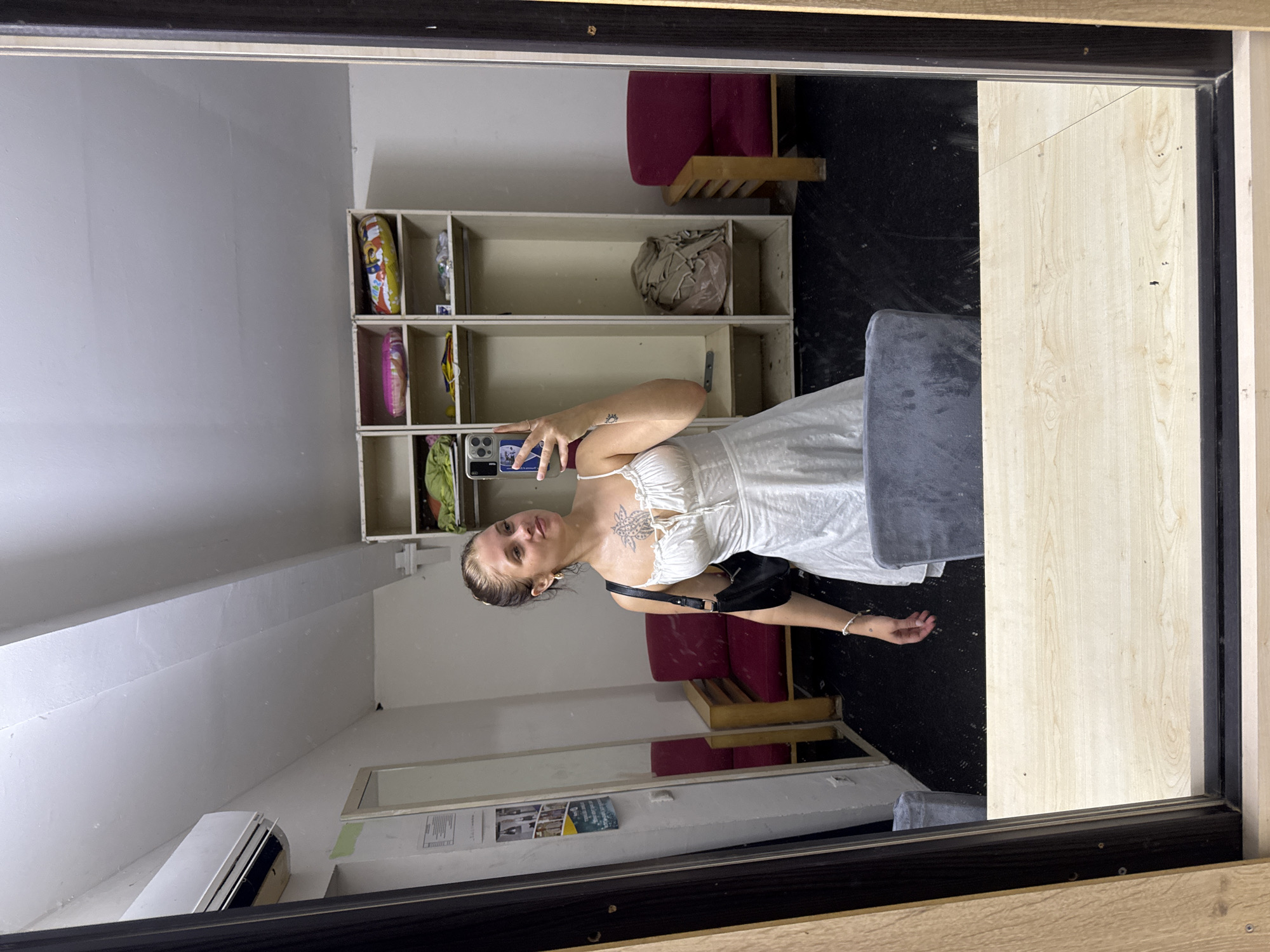 A young woman with blonde hair tied in a bun takes a mirror selfie. She is wearing a white, short, spaghetti-strap dress and a black shoulder bag. She has a tattoo on her chest and a small tattoo on her left arm. She is standing in a room with a black floor. To the left of the woman is a mirror reflecting the room, an air conditioning unit mounted high on the wall, and a poster on the wall. To the right of the woman, shelves are built into the wall, filled with various items including what appear to be balloons and folded clothes. Two red chairs with wooden frames are visible in the background, one to the left and one to the right of the shelves. A grey chair is partially visible in the foreground, in front of the woman. The image appears to be taken in Aksu, Turkey.