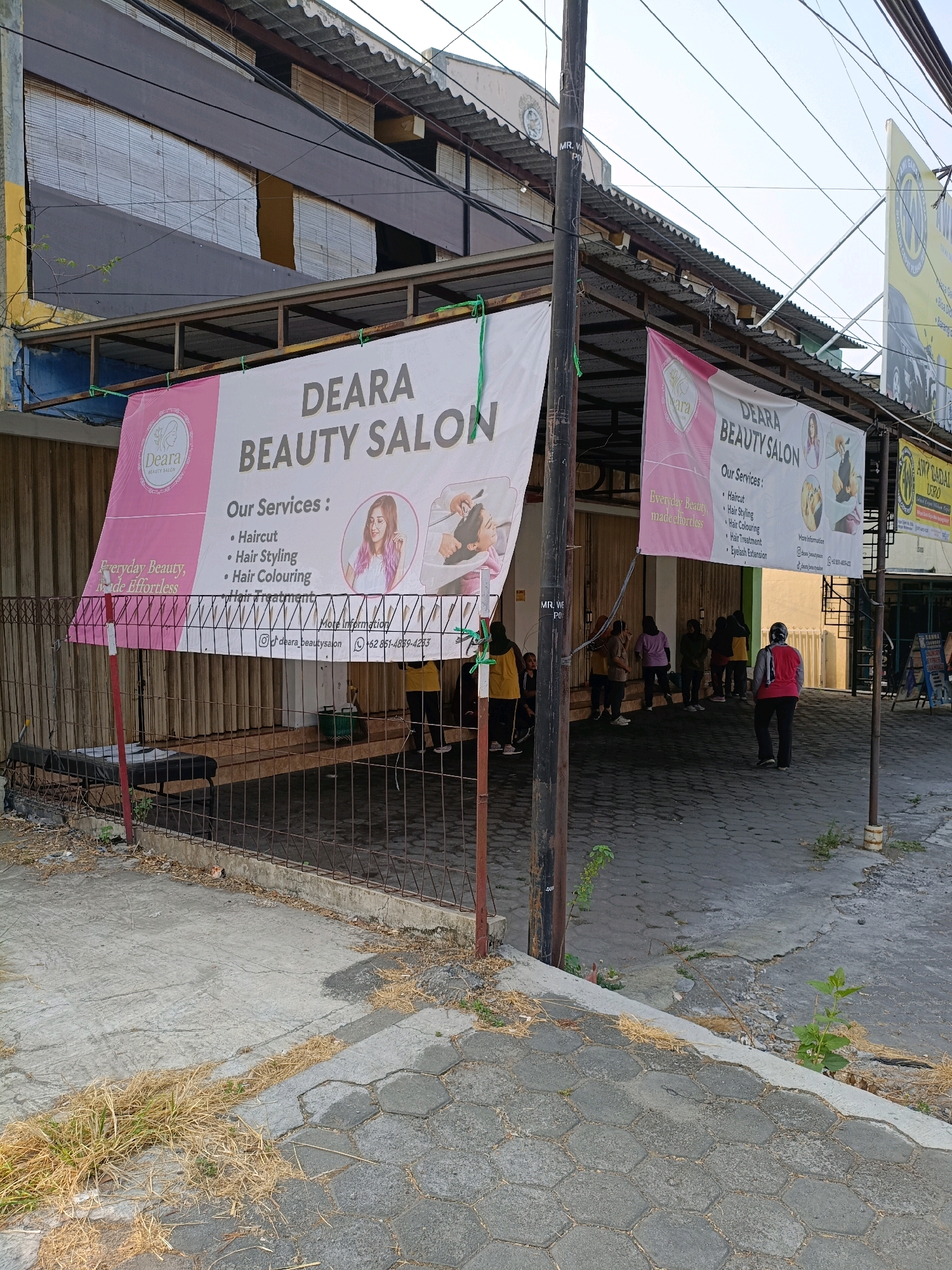The image shows the exterior of a beauty salon named "DEARA BEAUTY SALON" located in Ngemplak, Indonesia. Two banners are prominently displayed. The larger banner on the left features the salon's name, its logo, a slogan "Everyday Beauty, Made Effortless," and a list of services including "Haircut, Hair Styling, Hair Colouring, Hair Treatment, Eyelash Extension." Below this, contact information includes an Instagram handle "@deara_beautysalon" and a phone number "+62 851-4889-4253". A smaller banner on the right also displays the salon's name and services.
In front of the salon, there is a paved area with hexagonal stones, and a concrete walkway. A metal fence encloses an area with a massage bed visible inside. Several people are standing or walking in front of the salon, some appearing to be customers or staff. The building behind the banners has a corrugated metal roof and appears to be a commercial establishment. A utility pole with wires is positioned in the foreground. The overall environment is an urban street scene in daylight.