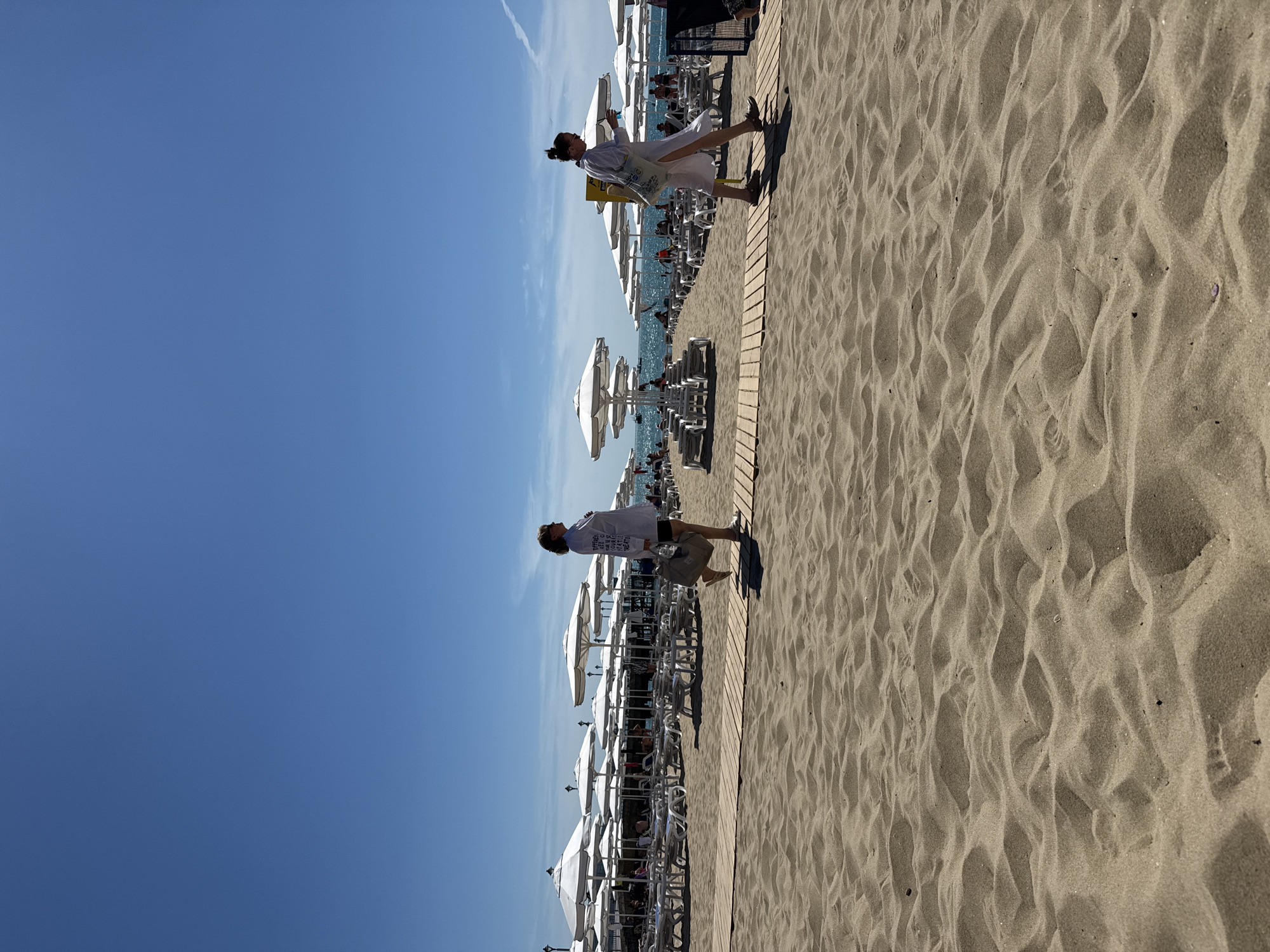 The image depicts a sunny day at a beach in Nesebar, Bulgaria. The foreground is dominated by sand dunes, with a wooden walkway extending into the background. On the walkway, two women are walking. The woman on the left wears a white shirt and shorts, carrying a large tote bag. The woman on the right is in a white dress and also carries a tote bag. In the background, numerous white beach umbrellas and lounge chairs are arranged neatly on the sand. The sea is visible beyond the beach furniture, with a clear blue sky overhead. The scene suggests a relaxing day at the resort.