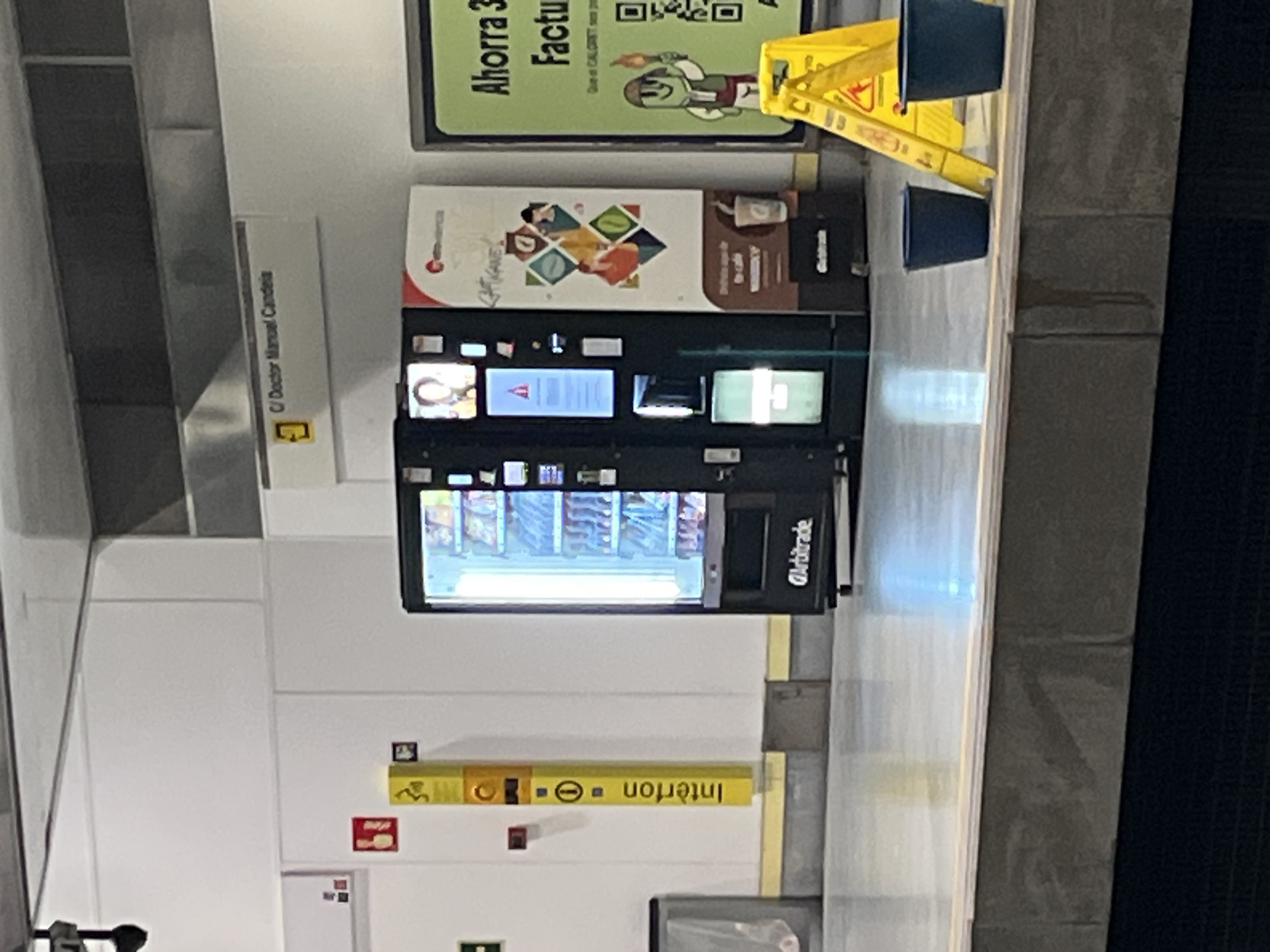 The image depicts an indoor scene in València, Spain. A vending machine prominently features the "Arbitrade" logo and is stocked with various items. The machine is located at the "C/ Doctor Manuel Candeia," as indicated by a nearby sign. Above the machine, a poster promotes savings on "Factu" purchases with "CALORET." The setting appears to be a public area, possibly a metro station, indicated by "Interfon" signage. A yellow "Wet Floor" sign is partially visible near the vending machine. The scene is well-lit, highlighting the vending machine and its surroundings.