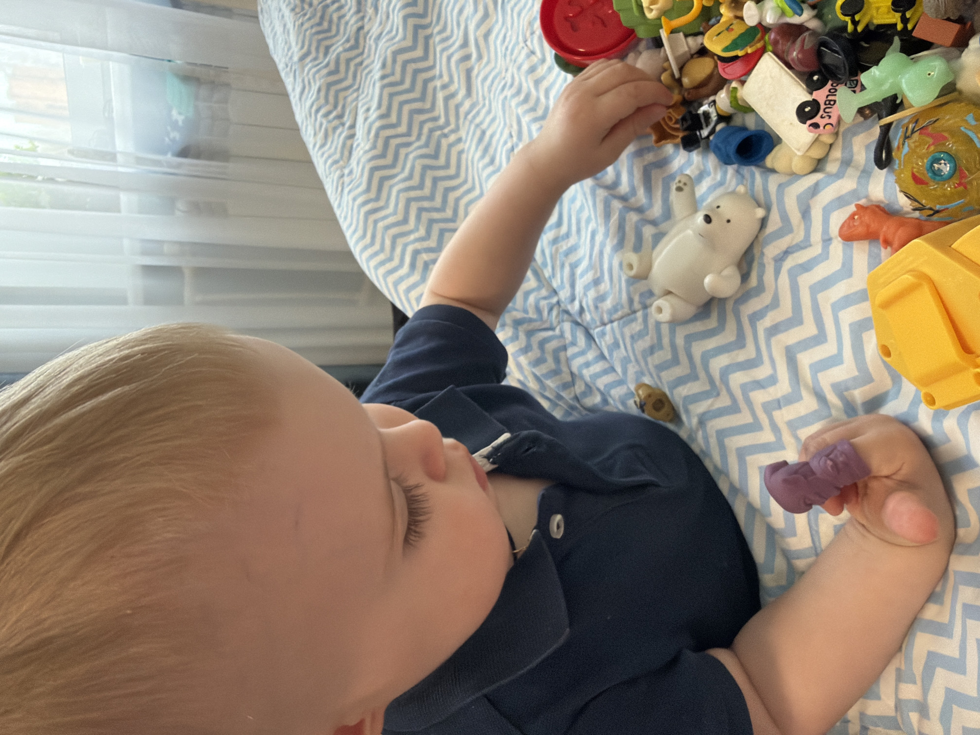 A child with blonde hair, wearing a dark blue shirt, lies on a bed covered with a blue and white zigzag patterned sheet. The child is surrounded by various toys. The exact location within the city of Votkinsk, Russia, cannot be determined from the image.