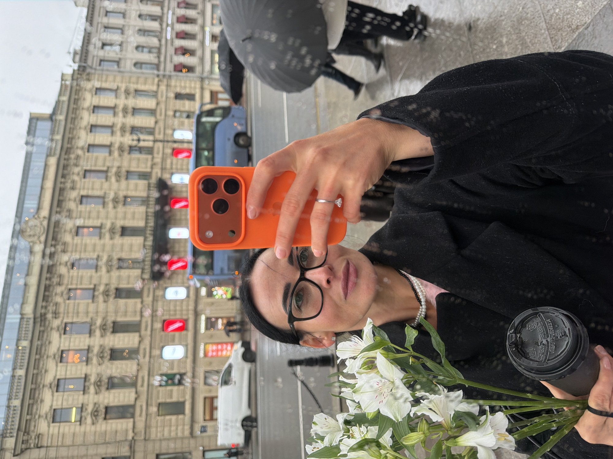A woman with black hair and glasses holds an orange smartphone, a bouquet of white alstroemeria flowers, and a black coffee cup while taking a selfie. The background shows a blurred city street with a beige building, cars, and pedestrians with umbrellas. The location is Saint Petersburg, Russia. The reflection on the glass suggests it is raining. The woman is wearing a dark coat and a pearl necklace. She is holding the phone in her left hand. A silver ring is visible on her finger.