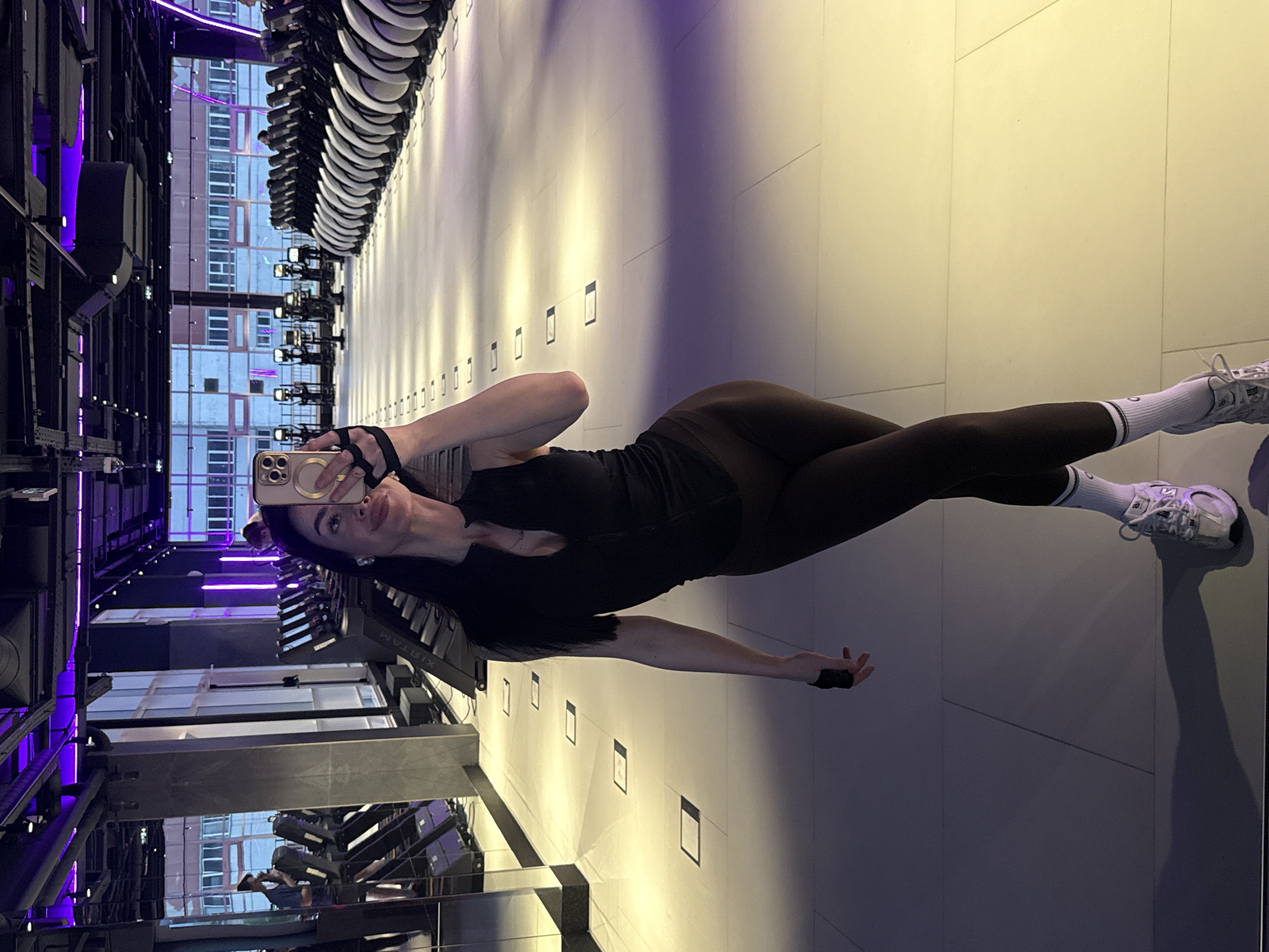A woman in black leggings, a black top, white socks with black stripes, and white sneakers lies on her side on a tiled floor. She holds a smartphone in her left hand, taking a selfie. The background reveals an interior space with rows of exercise equipment under fluorescent lighting, and a view of a building exterior through a window. The equipment appears to be rowing machines, arranged in a line along a wall. Based on the "XISTAM" on the equipment, the location appears to be XISTAM FIT located in Krasnoyarsk, Russia.
