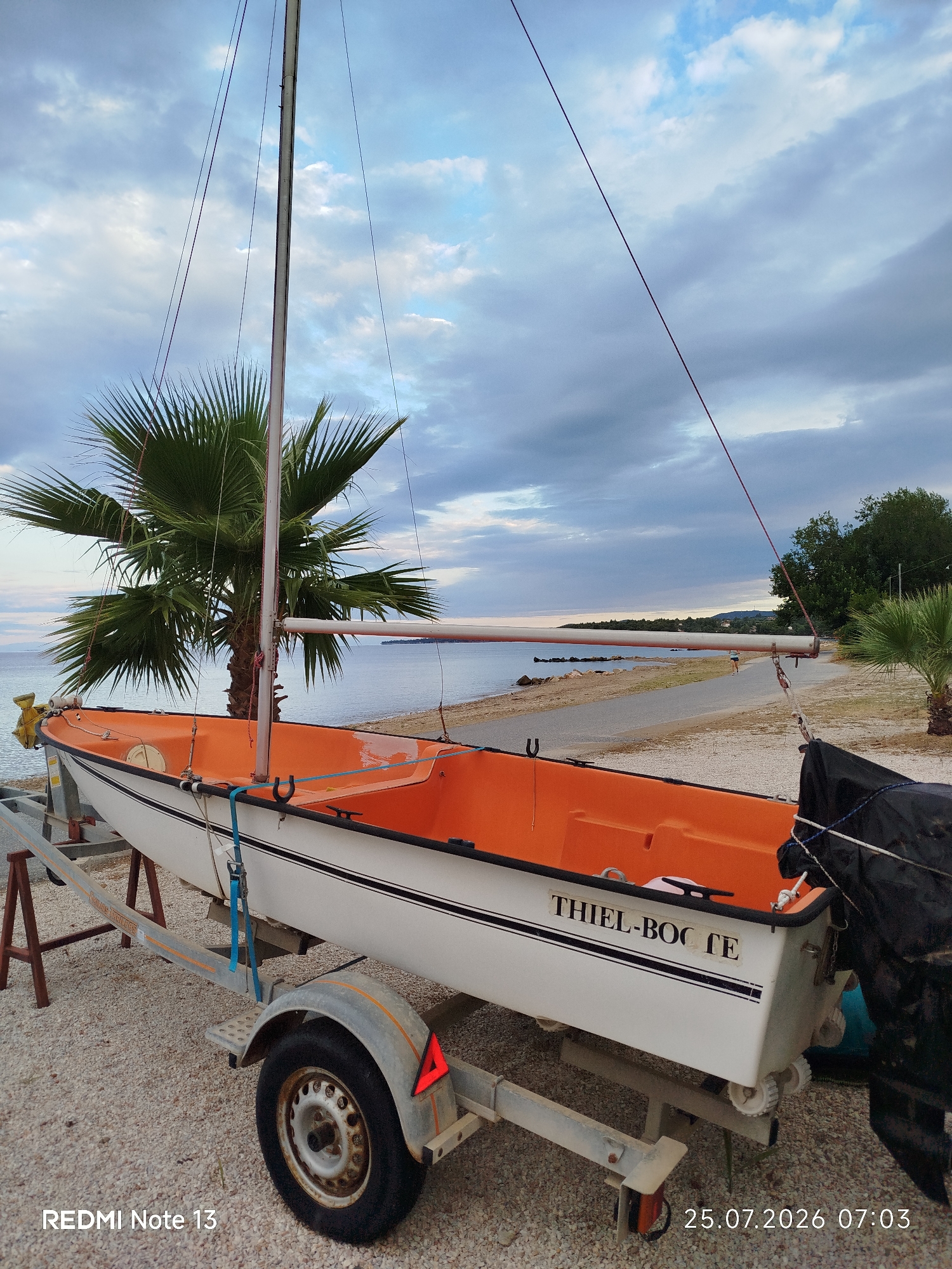 This image depicts a small sailboat with an orange interior, resting on a trailer on a pebble beach. A palm tree is visible behind the sailboat, with the sea and a distant shoreline in the background. The sky is partly cloudy. The sailboat has "THIEL-BOOTE" written on its side. The location is identified as Sithonia Municipal Unit, Greece, with the timestamp 25.07.2026 07:03 and "REDMI Note 13" visible in the bottom left and right corners respectively.
