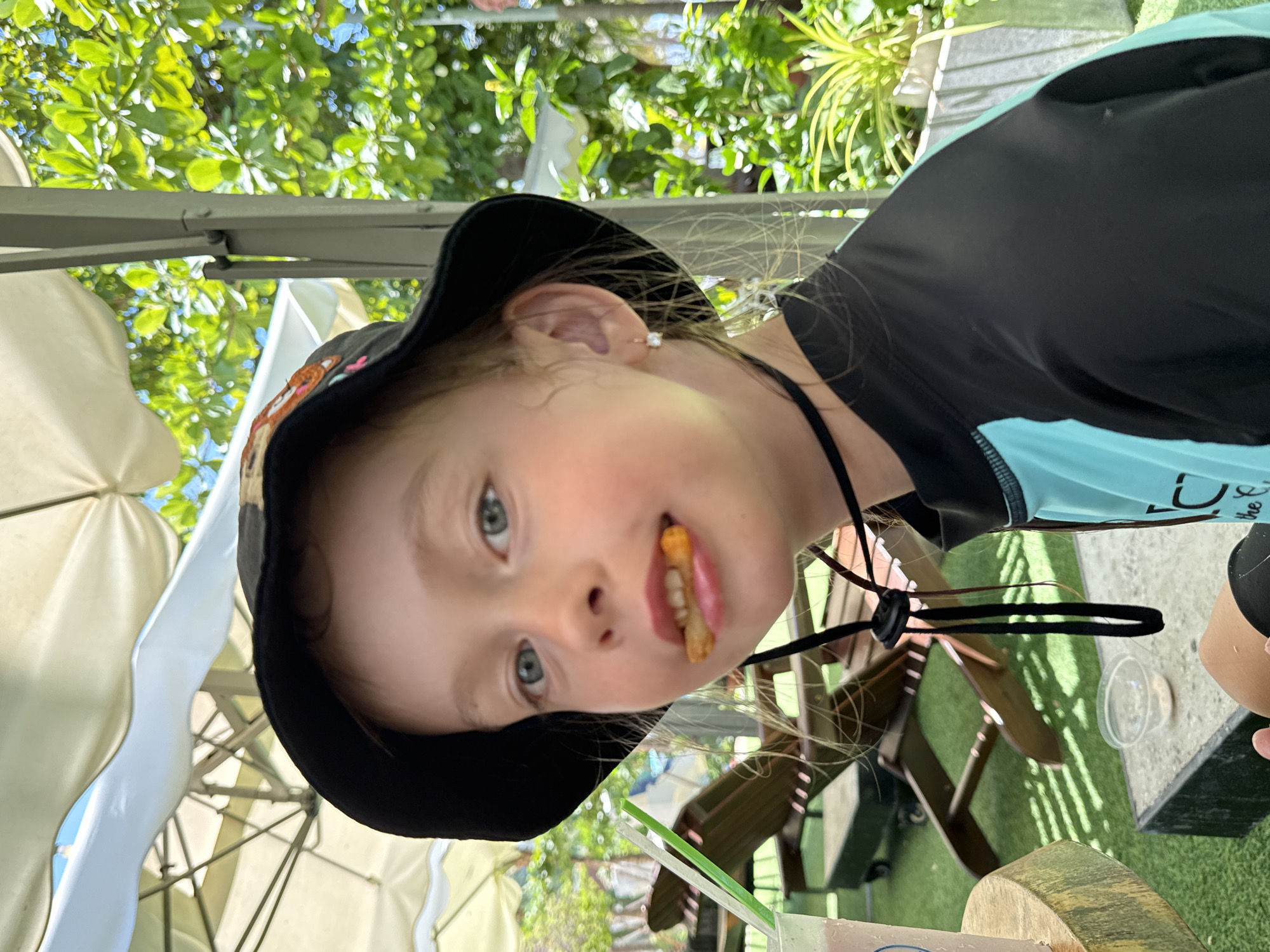In Nha Trang, Vietnam, a young Caucasian girl with light brown hair, wearing a black bucket hat with an embroidered patch and a turquoise and black rash guard, is captured in a close-up portrait. She is sitting outdoors, possibly at a cafe or restaurant, with a drink and straws visible in the foreground. A french fry is partially visible in her mouth. The background suggests an outdoor seating area with lounge chairs and lush green foliage, characteristic of a tropical environment. The lighting indicates it is daytime.