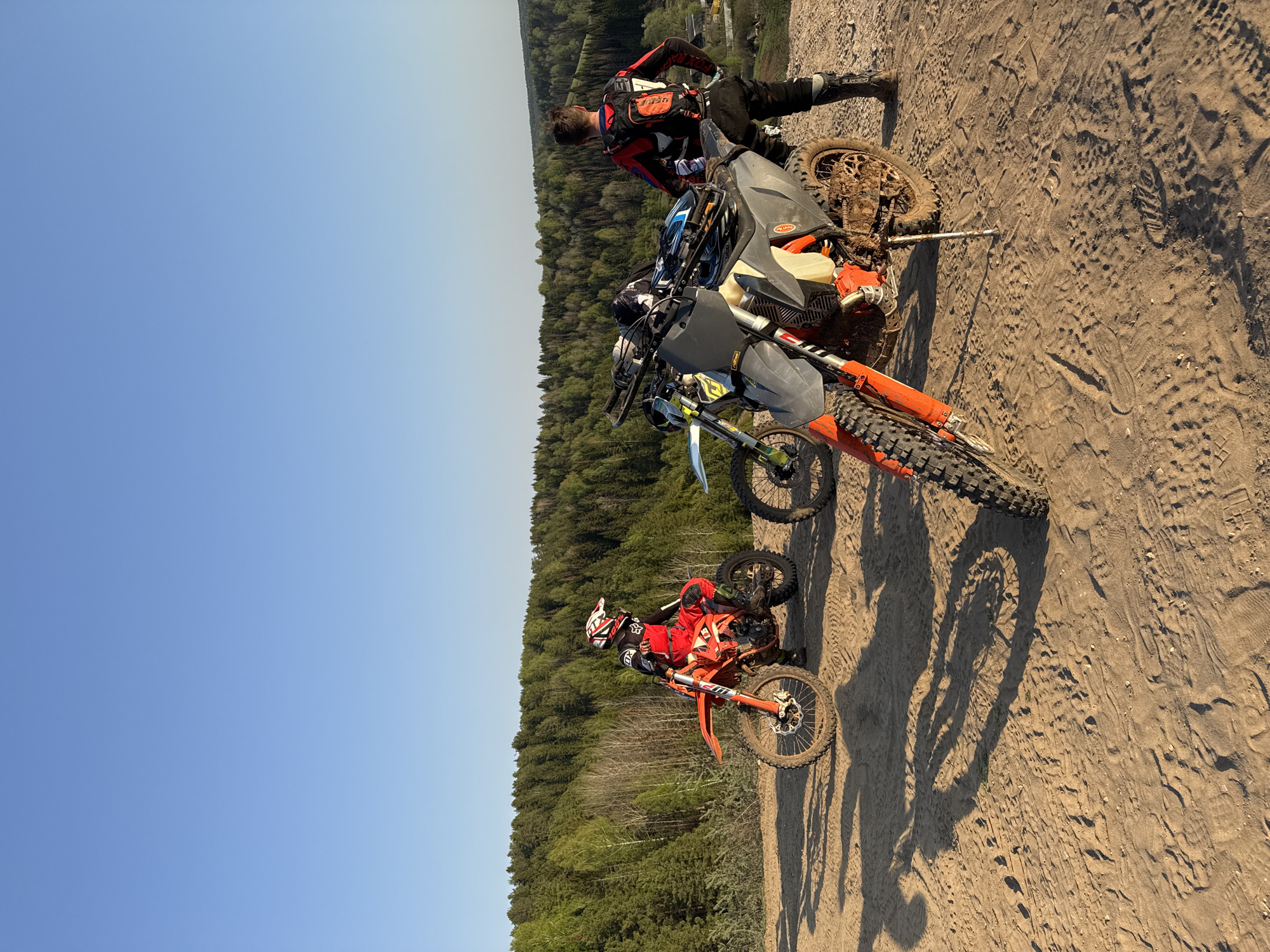 The image depicts a group of dirt bikers and their motorcycles in what appears to be a sandy or dirt terrain near a forest, likely in a rural area. The setting is in the vicinity of Ольховка, Russia, as specified.
Subjects:
- Three dirt bikes are visible, with at least one rider on each or nearby. The riders are wearing protective gear like helmets, goggles, and riding suits.
- One individual is standing near a bike.
Objects:
- The main objects are the dirt bikes, which vary in color and model.
- Riding gear (helmets, goggles, suits)
Actions:
- The individuals are posing for a picture and possibly resting or socializing, as the bikes are parked.
Environment:
- The ground is covered in sand or dirt with tire tracks and footprints.
- A forest is in the background.
- The sky is clear and blue.