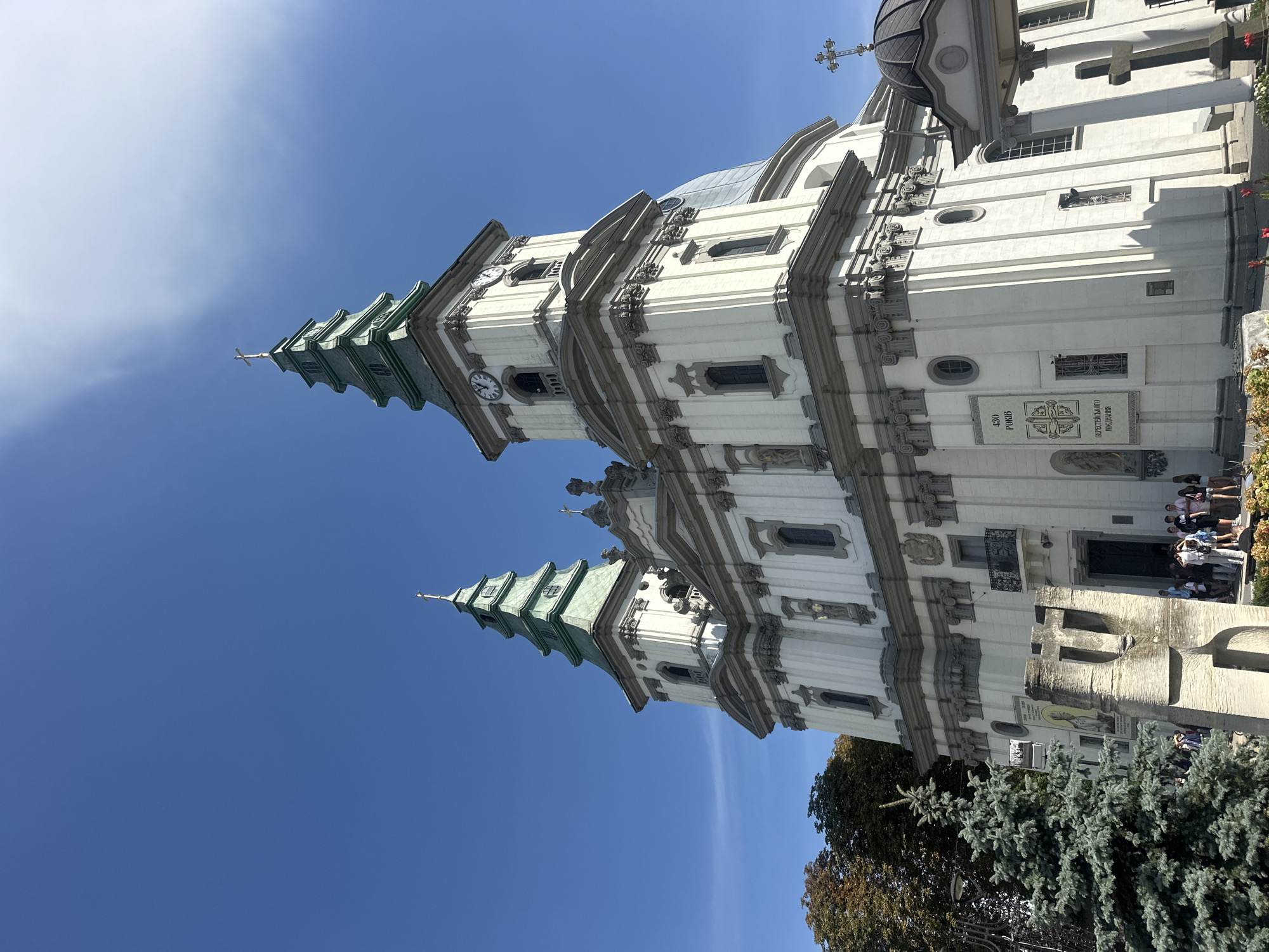 The image shows a large, white church with two green spires. The church has a classical architectural style with columns and ornate detailing. There are several windows and balconies on the facade. In front of the church, there is a group of people standing, some appearing to be tourists. A modern sculpture is also visible in the foreground, as well as a Christmas tree. The sky is a clear, bright blue with some wispy clouds. The church is likely the Church of the Immaculate Conception of the Blessed Virgin Mary in Ternopil, Ukraine, a prominent landmark known for its baroque architecture.