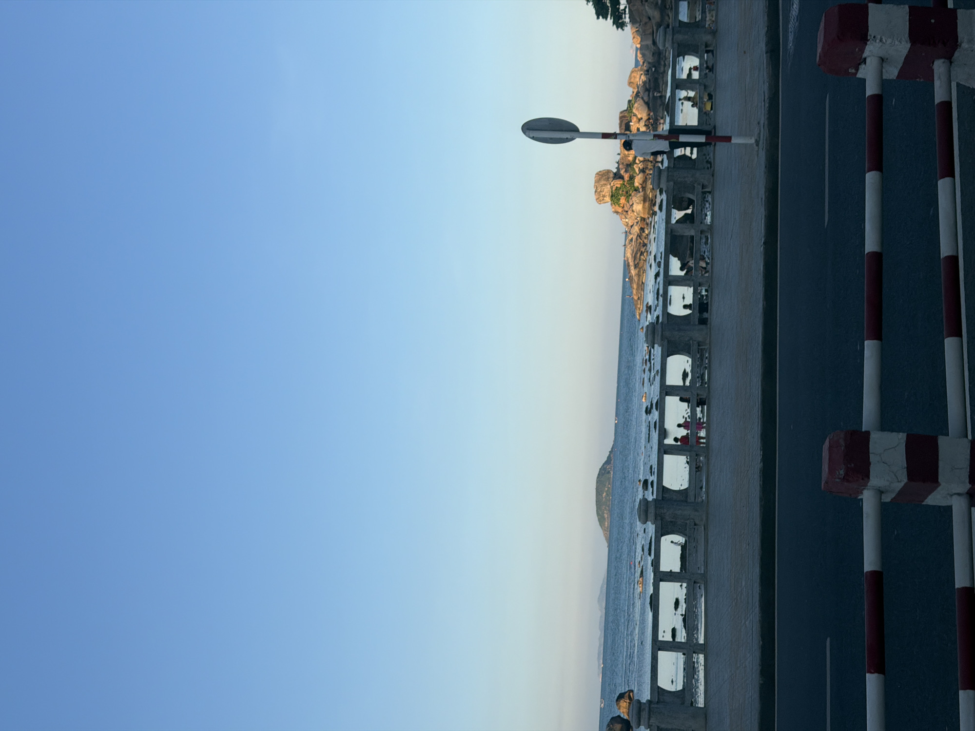The image shows a view of the ocean from a coastal road in Bac Nha Trang Ward, Vietnam. The sky is clear and blue. In the distance, there is a small island with vegetation. Along the coastline, there are rocky formations and the sea. A stone railing with square openings runs along the edge of the promenade. Several people are visible on the promenade, including two children standing near the railing and other figures in the background. A traffic signpost with red and white markings stands on the right side of the image. The foreground features a dark asphalt road with a red and white striped barrier. The overall scene is a peaceful coastal landscape.