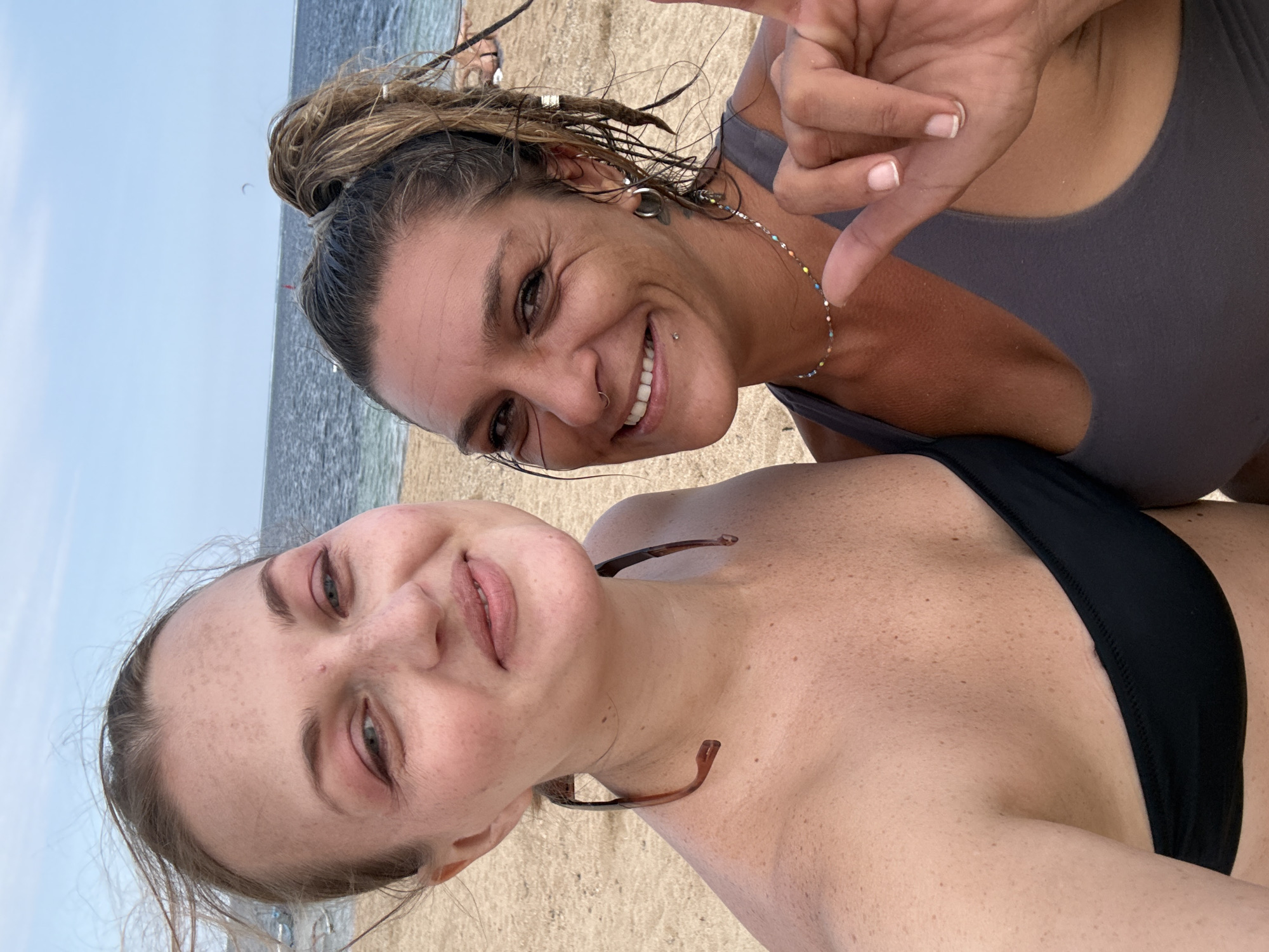 The image shows two women on a sandy beach with the ocean in the background. The woman on the left has blonde hair and is wearing a black bikini top. She has sunglasses hanging from her neck. The woman on the right has brown hair pulled back in a ponytail and is wearing a grey tank top. She is making a hand gesture. The sky is partly cloudy. There is no indication that this image was taken in Denpasar, Indonesia.