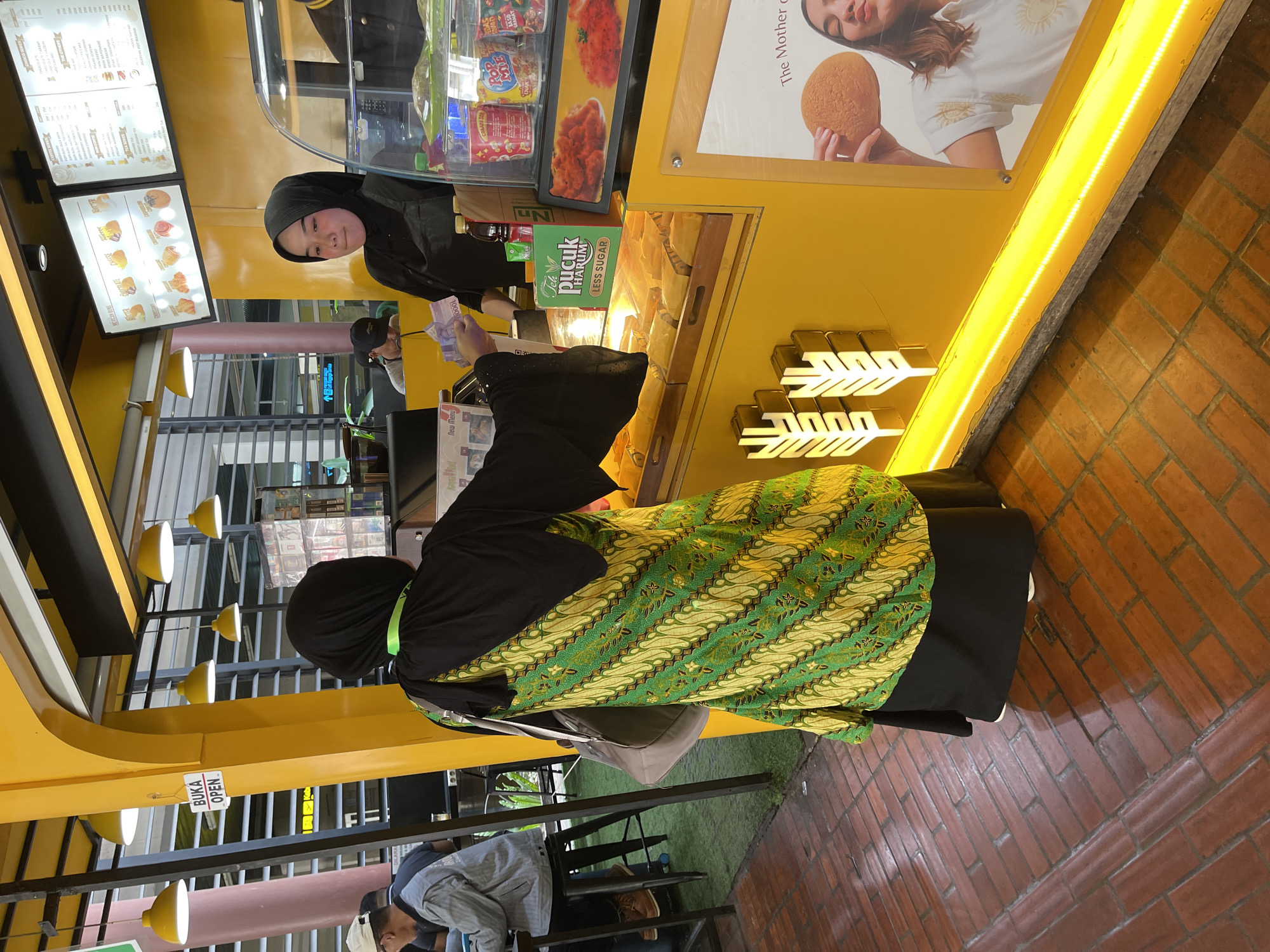 The image depicts an outdoor food stall with a bright yellow facade, illuminated by yellow lights. Two individuals are present. In the foreground, a person wearing a black headscarf and a vibrant green and yellow batik-patterned garment is standing with their back to the camera, holding money and interacting with a vendor. The vendor, also wearing a black headscarf and an apron, is behind the counter, looking down and appears to be engaged in a transaction. The counter displays various food items, including pastries, and packaged drinks such as "Teh Pucuk Harum". In the background, another person is seated at a table. A sign displaying "BUKA OPEN" is visible on a yellow structure. The flooring is made of red brick. The presence of the "Teh Pucuk Harum" product, a widely available beverage in Indonesia, and the general aesthetic suggest the location is likely within a public area or mall in Tangerang, Indonesia.