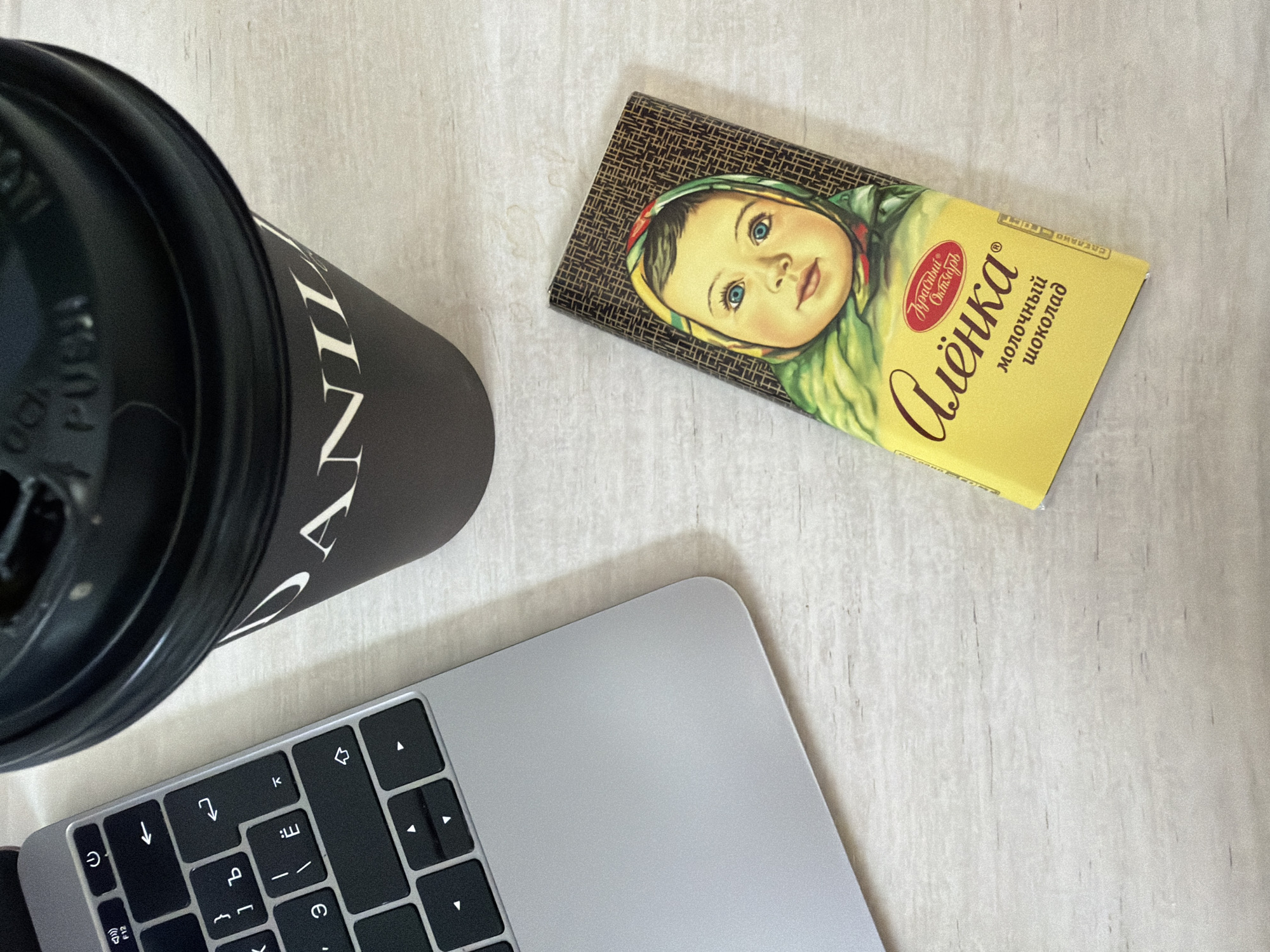 This top-down, slightly angled shot captures a casual work or break scene on a light-colored, textured surface, possibly a wooden desk or countertop, in Chaikovsky, Russia.
In the upper left, a black coffee cup or thermos with a lid is partially visible. Text on the side of the cup shows "ANIL" which is likely part of "DANIEL" or "DANIELL'S," and the word "PUSH" is clearly visible on the lid.
Dominating the upper right is a bar of "Alyonka" milk chocolate, a famous Russian brand, which reinforces the given location context. The wrapper features a classic illustration of a baby's face wearing a headscarf. Visible Russian text on the wrapper includes "Красивый Октябрь" (Krasivyy Oktyabr, meaning 'Beautiful October'), "Алёнка" (Alyonka), and "молочный шоколад" (molochnyy shokolad, meaning 'milk chocolate'). Further text "СДЕЛАНО ГОСТ" (SDELANO GOST, meaning 'Made to GOST standard') is partially visible on the right edge.
In the lower left, a silver laptop, likely a MacBook due to its design, is open, showing a portion of its black keyboard. The keys display a mix of Cyrillic and Latin characters, along with arrow keys and other function keys.
The scene suggests an indoor setting, likely during the day, with soft, even lighting, indicative of a typical home or office environment. No specific activity is captured beyond the arrangement of these items, hinting at a moment of rest or focused work.