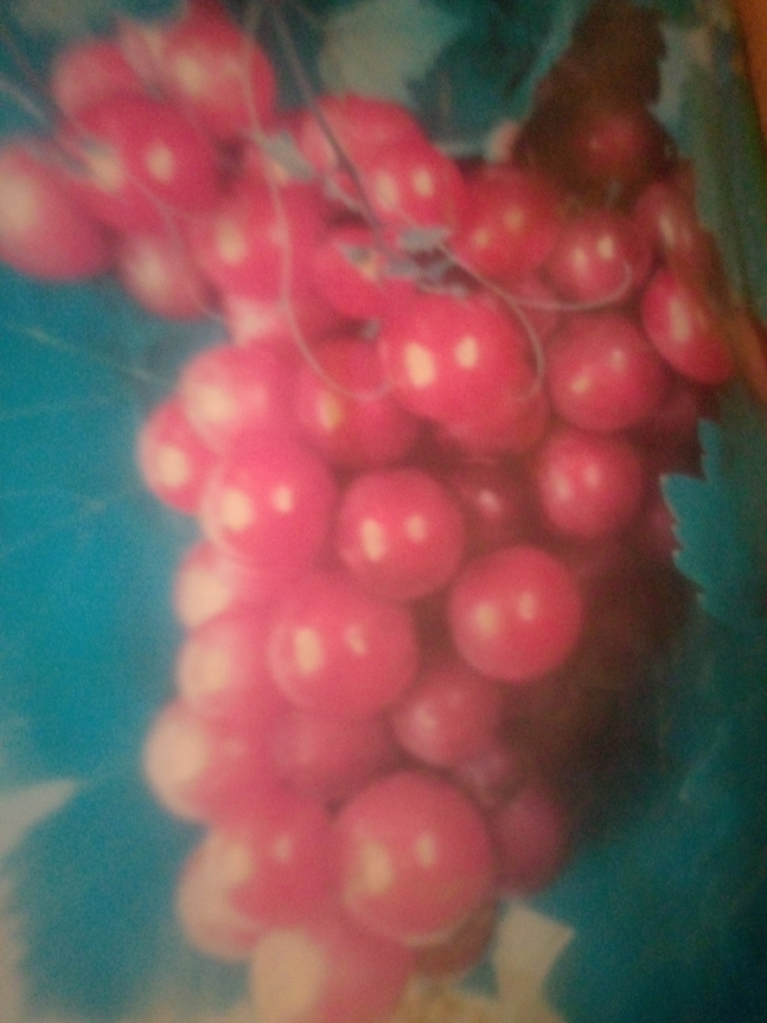 The image depicts a close-up of a bunch of pink grapes against a blurred turquoise background. The grapes are round and plump, with some appearing to have a slight sheen, suggesting they are ripe. White highlights on the surface of the grapes indicate points where light is reflecting. The overall impression is one of abundance and freshness. The background is a solid, vibrant turquoise, which contrasts with the pink of the grapes, making them stand out. There is a hint of darker green foliage visible on the right side of the image, suggesting the grapes are still on the vine or have recently been picked. The focus is entirely on the grapes, with the background out of focus to draw attention to the main subject. There is no visible text in the image. The image appears to be a still photograph, possibly taken in natural light given the highlights on the grapes. No people are visible.