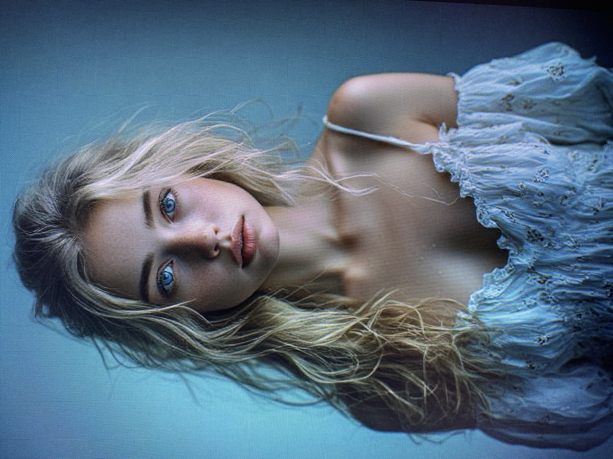 The image is a close-up, vertically oriented photograph of a young woman lying down. She has long, blonde, wavy hair that is spread out around her head and shoulders. Her eyes are a striking blue and are looking towards the camera. Her lips are slightly parted. She is wearing a light blue, off-the-shoulder top with delicate white lace trim and a floral pattern. The background is a soft, solid blue, creating a minimalist and serene setting. The lighting appears soft, illuminating her face and the textures of her hair and clothing. The overall mood is one of peacefulness and contemplative beauty. There is no visible text in the image. The context of Yerevan, Armenia is noted but does not provide specific visual cues within the image itself.