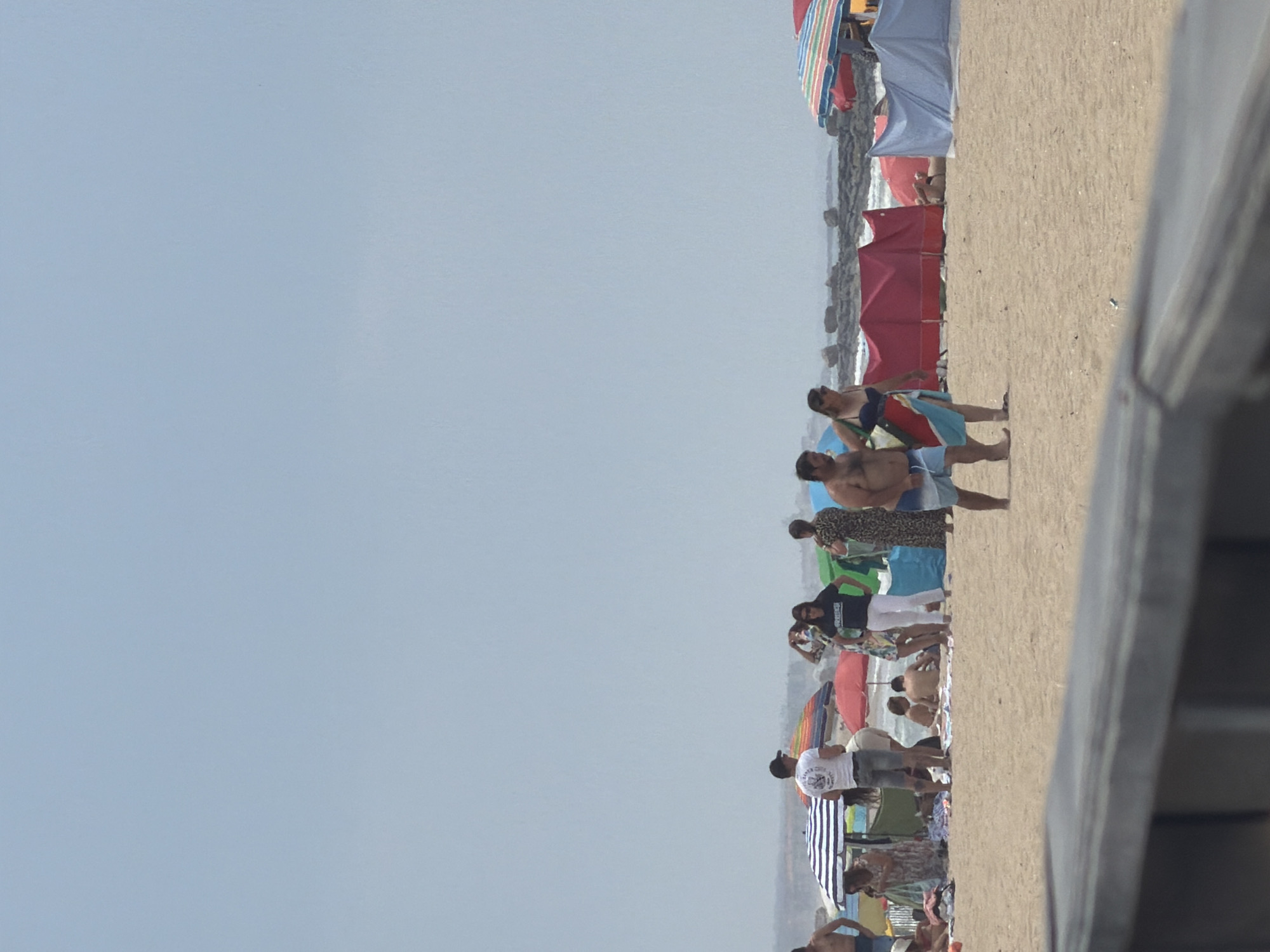 The image depicts a crowded beach scene on a sunny day, likely in Espinho, Portugal, given the context. The foreground is dominated by a wooden railing, obscuring the lower portion of the beach. The sandy beach is populated with numerous individuals, some in swimwear, others in casual clothing. Various beach umbrellas, including striped and solid colored ones, are scattered across the sand, providing shade. Several beach shelters, some red and some blue, are set up, with people visible inside or near them. In the mid-ground, a couple walks hand-in-hand, the man wearing blue shorts and the woman in a blue bikini top and a colorful sarong. Other people are seen standing, sitting, and interacting amongst themselves. The background shows the ocean with waves breaking against rocky outcrops. The sky is a pale, hazy blue.