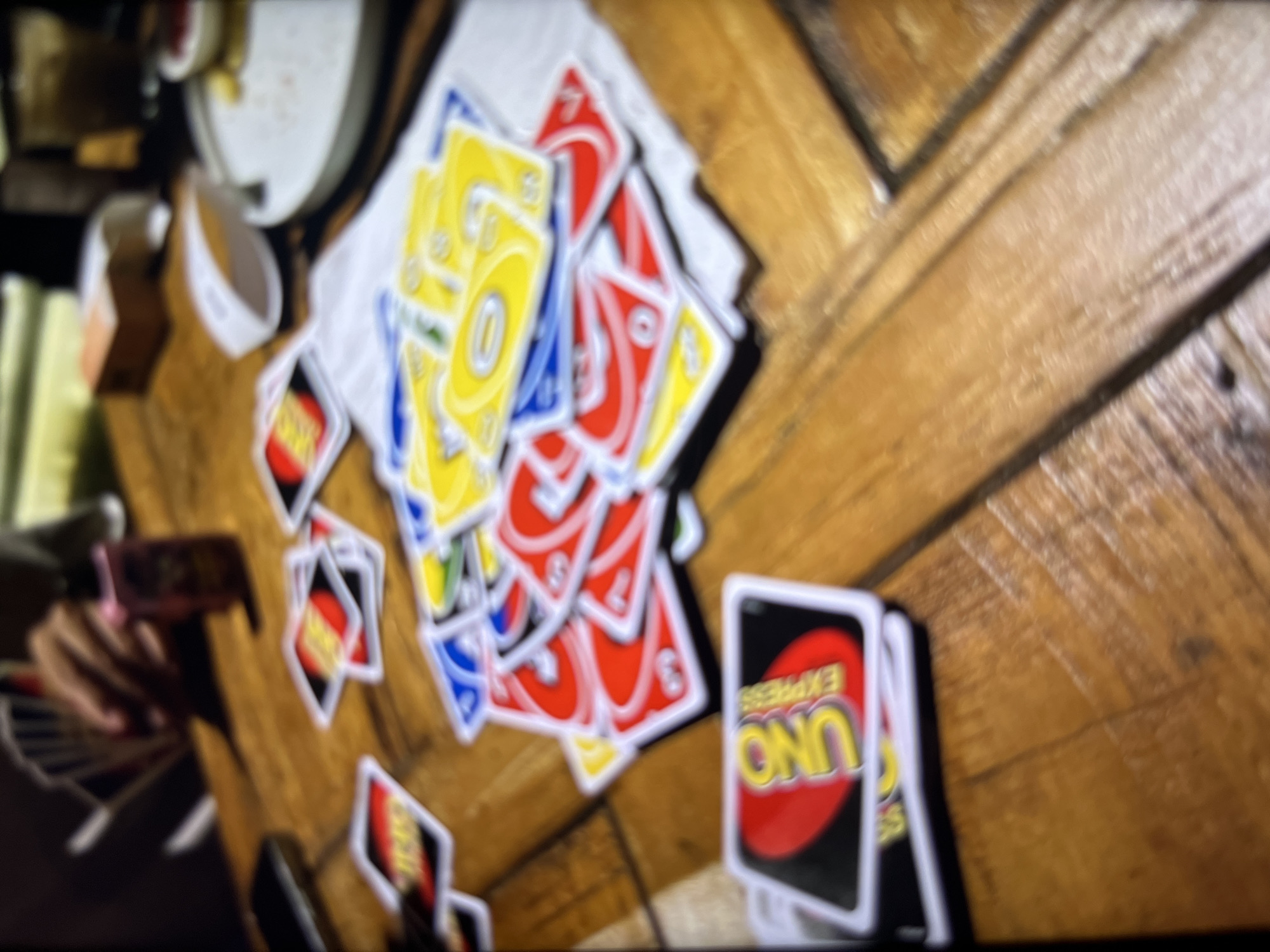 A pile of UNO cards is scattered across a wooden table. In the background, a person's hand is visible, holding a fan of cards. There are also other objects on the table, including what appears to be a white electronic device, a plate with food, and a box. The lighting suggests it is daytime or a well-lit indoor setting. The overall impression is a casual gathering or game in progress. The location is stated as Bukuru, Nigeria.