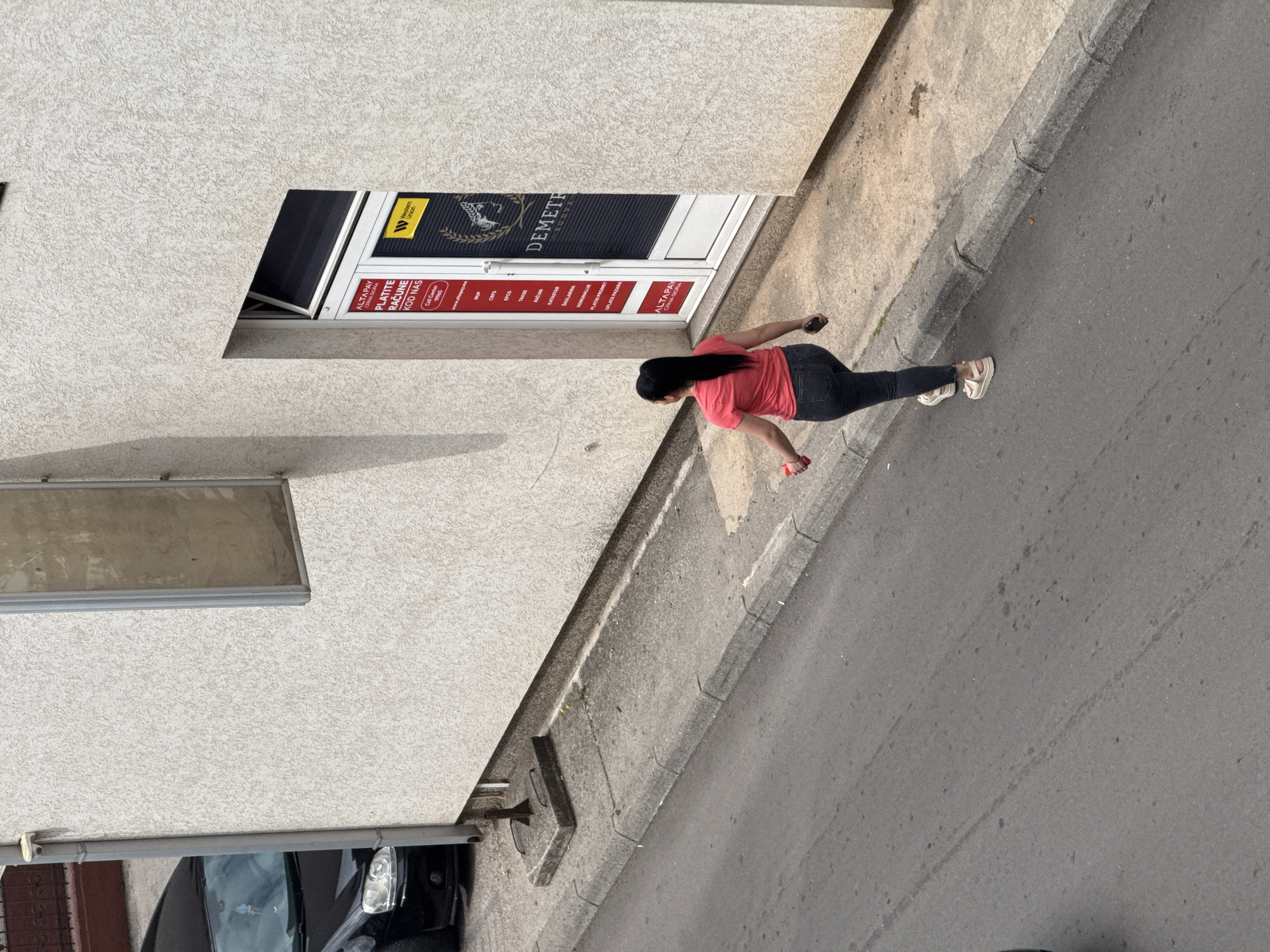 The image captures an elevated view of an urban street scene in Budva, Montenegro, during clear daytime weather. A woman with long dark hair, dressed in a red short-sleeved top, dark jeans, and light-colored platform sandals, is walking along the asphalt road next to a building. She appears to be holding a small red object in one hand and possibly a mobile phone in the other.
The building features a light-colored, textured facade. A shop entrance is visible, identified by a dark door with signage. Prominent text includes "Western Union" and "DEMETH BUDVA." Below this, a red and white sign displays services offered by "ALTAPAY CRNA GORA," with the instruction "PLATITE RAČUNE KOD NAS" (Pay Bills With Us). Specific services listed are "MUP," "EPCG," "TAKSI," "RAČUNI," "KATASTAR," "ŠKOLARINA," "KOMUNALIJE," "PLATNI PROMET," and "UPLATA PAZARA." Contact details "Call Center 19960" and "www.altapay.me" are also present.
A grey concrete curb separates the building from the street. The asphalt road shows some minor marks and stains. In the bottom left, part of a dark car is visible, parked beneath a structure attached to the building.