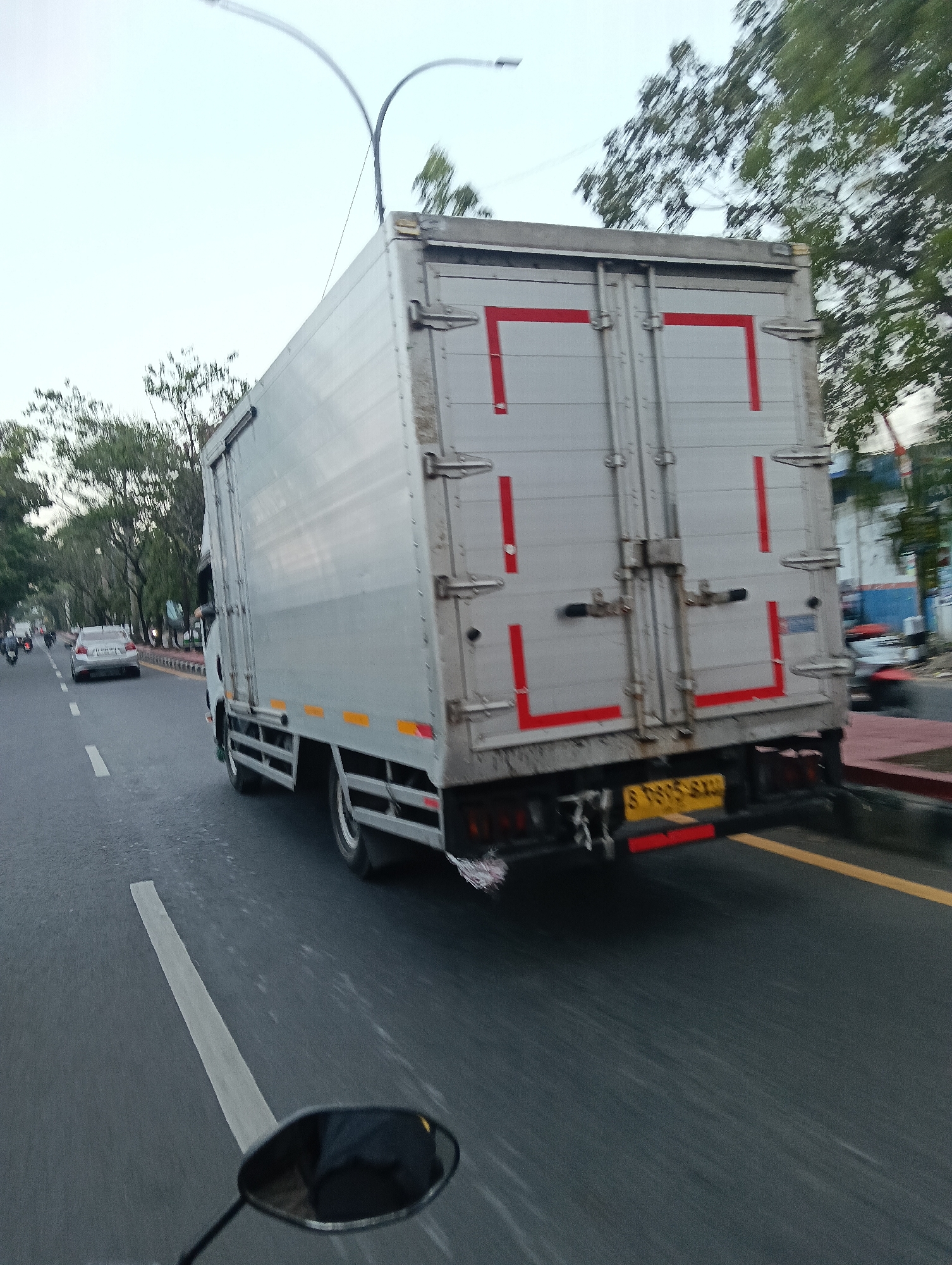 The image shows a street scene in Tasikmalaya, Indonesia, likely during the day with overcast skies. A large, silver box truck is the dominant subject, seen from its rear and right side. The truck has red markings on its rear doors and yellow license plates. It appears to be moving on a paved road with white lane markings.
To the left of the truck, a silver car is visible, also in motion. Further down the road, several motorcycles are present. On the right side of the road, a sidewalk is visible with some trees lining it. In the background, there are buildings and more trees, indicating an urban or semi-urban environment. The perspective suggests the image was taken from a moving vehicle, possibly a motorcycle, as a portion of a rearview mirror is visible in the foreground. The overall impression is of traffic on a road in Tasikmalaya.